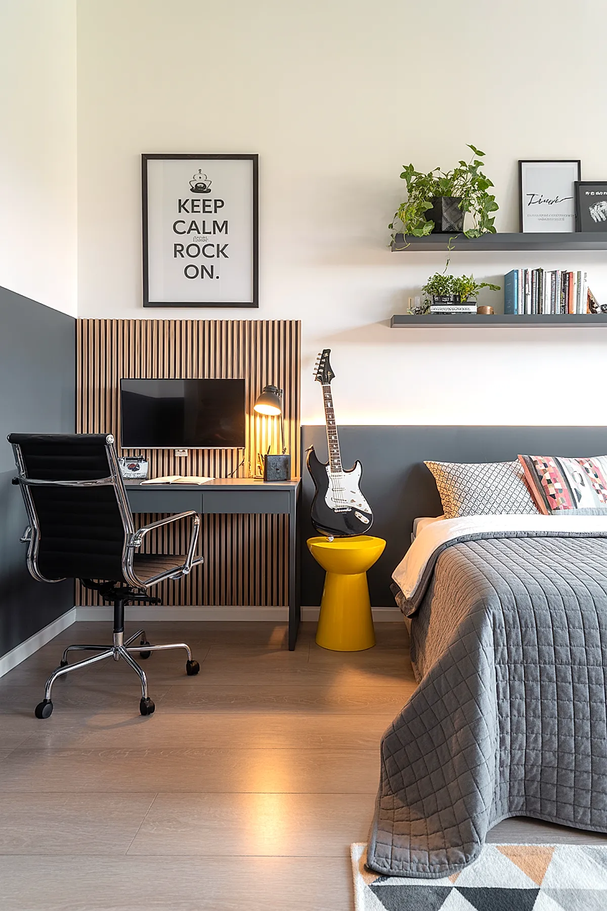 Teen boy bedroom featuring gray and white walls, wooden slat accent behind desk, black ergonomic chair, small gray desk with lamp and computer monitor, black electric guitar resting on bright yellow stool next to bed with gray quilted bedding, floating shelves with plants and books above.