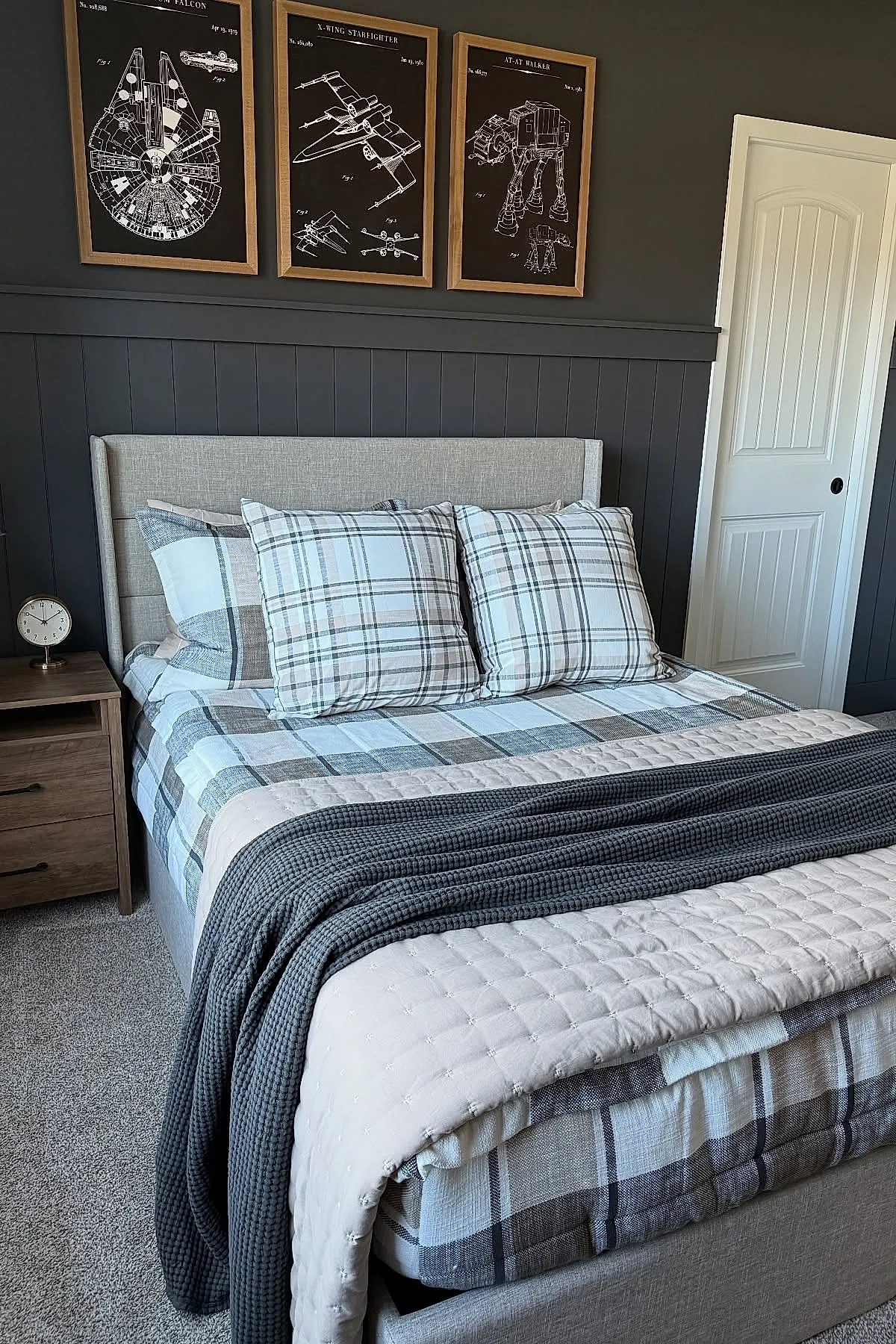 Teen boy bedroom with dark gray vertical paneled wall behind bed, beige upholstered headboard, bedding in muted plaid patterns of gray, white, and beige. Above bed are three wooden framed black and white Star Wars vehicle schematics hung evenly. Light wood nightstand with small round clock sits to left.