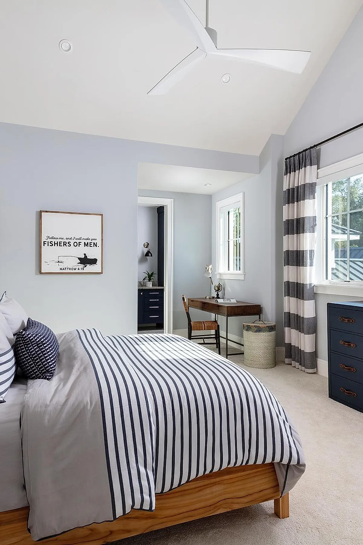 Teen boy bedroom with light gray walls, wooden bed frame with white and navy narrow striped bedding, navy and white wide striped curtains, wooden desk with chair near windows, beige carpet floor.