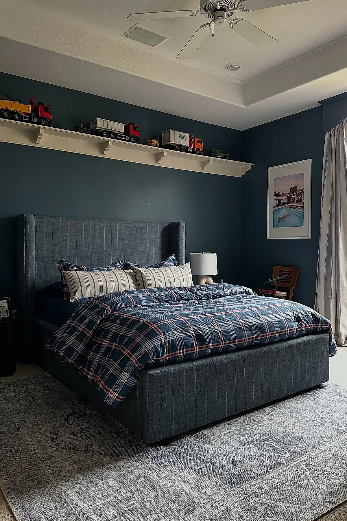 Teen boy bedroom with dark blue walls and matching upholstered bed frame, plaid blue bedding, white ceiling fan, long narrow white shelf near ceiling holding toy trucks, framed picture on adjacent wall, and light gray patterned area rug.