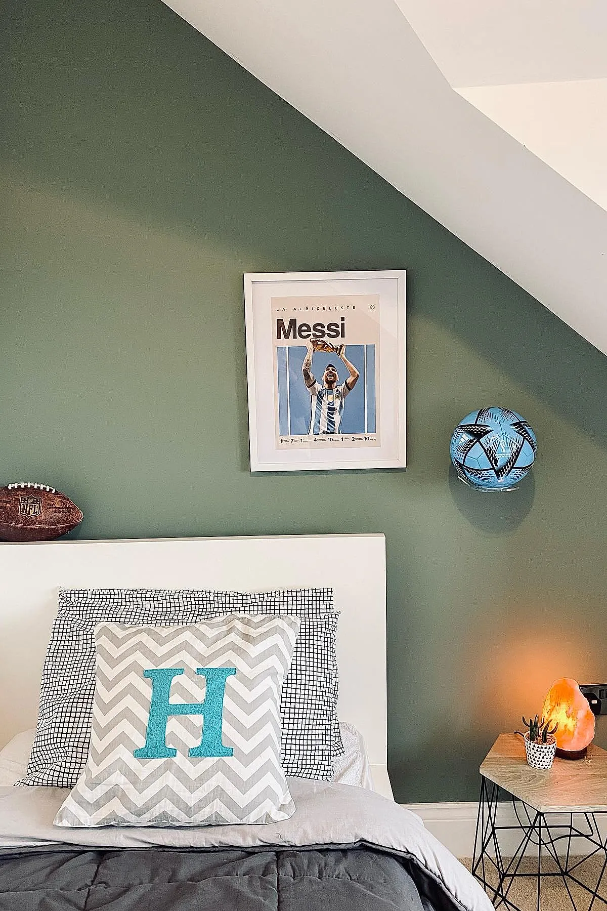 Teen boy bedroom featuring a white bed frame against an olive green accent wall, chevron patterned pillow with large turquoise letter H, black and white grid pillowcases, framed Messi poster above bed, brown NFL football on headboard edge, blue soccer ball mounted on wall, wooden side table with small plant and Himalayan salt lamp.