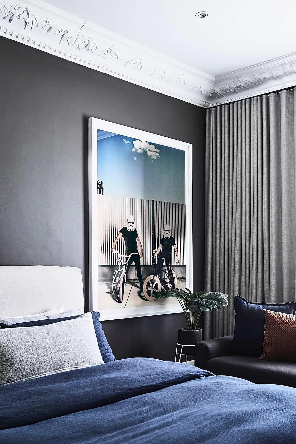Teen boy bedroom with dark gray walls, large framed photo of two people on bikes wearing stormtrooper helmets, white upholstered bed with blue bedding, gray curtains, black armchair with navy and rust pillows, and a small plant on a black stand.