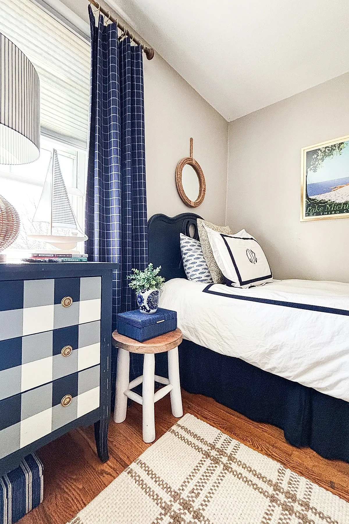 Teen boy bedroom featuring a navy blue checkered curtain, matching checkered dresser in shades of blue and white with brass knobs, white bed linens with navy trim, patterned pillows, small wooden stool with plant in blue and white ceramic pot, rope-framed round mirror on beige wall, and framed Lake Michigan poster.