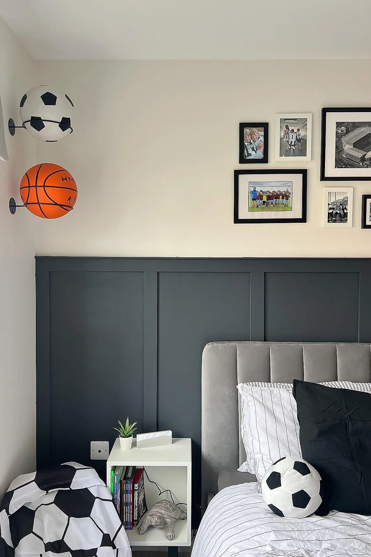 Teen boy bedroom corner featuring two sports balls mounted on the wall-a basketball and a soccer ball-above a white nightstand with books and a small plant, gray upholstered bed with striped bedding, and framed sports photos on the wall.