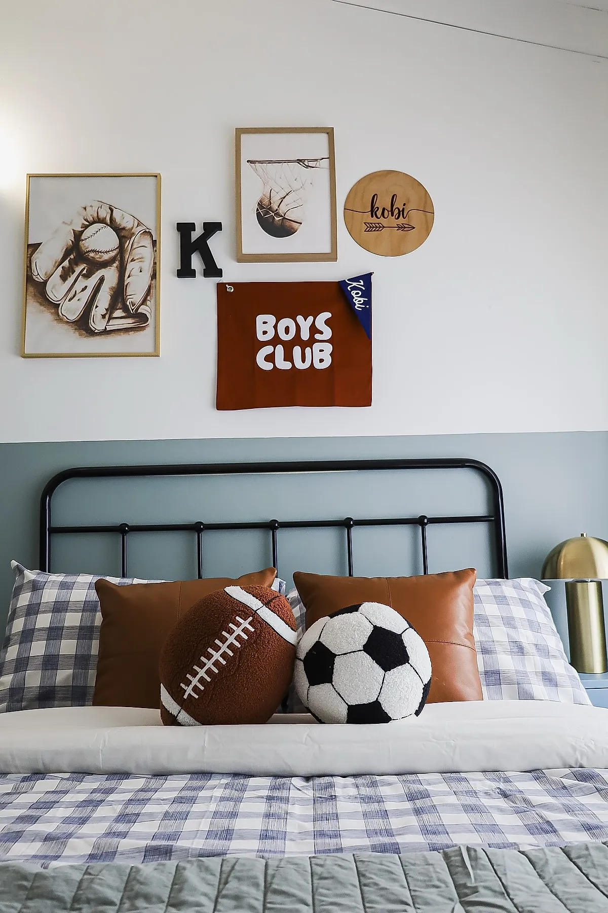 Teen boy bedroom with black metal bed frame, blue and white checkered bedding, brown leather pillows, plush football and soccer ball pillows at center, gallery wall featuring framed baseball glove print, basketball hoop print, wooden round sign with name Kobi, black letter 'K', and brown Boys Club pennant.
