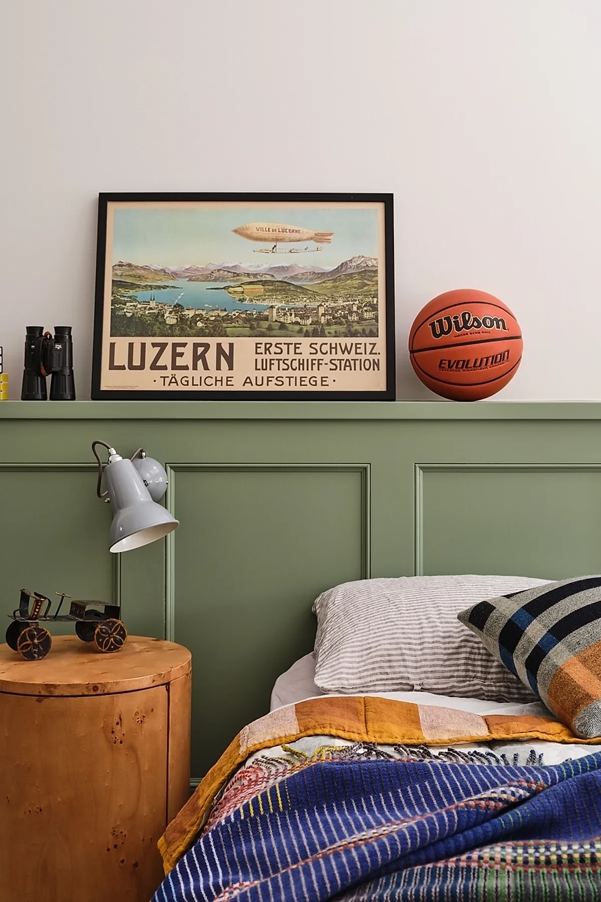 Teen boy bedroom with muted green paneled wall, framed vintage Luzern travel poster resting on the ledge above bed, an orange Wilson basketball placed next to it, gray desk lamp attached to the side of the ledge, round wooden side table with small metal car model, striped bedding in blue, mustard yellow, and white tones.