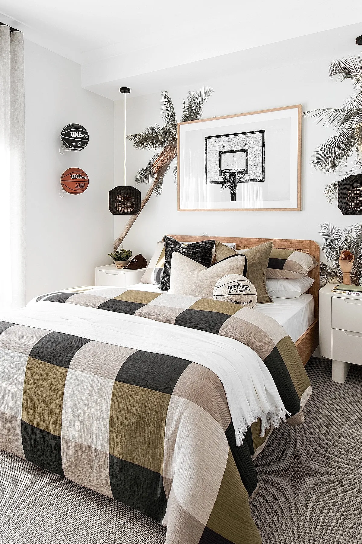 Teen boy bedroom featuring a bed with plaid bedding in beige, black, olive, and cream tones; large framed black-and-white basketball hoop photo above headboard; palm tree wall mural behind bed; two basketballs mounted vertically on white wall; light beige carpet and white bedside tables.