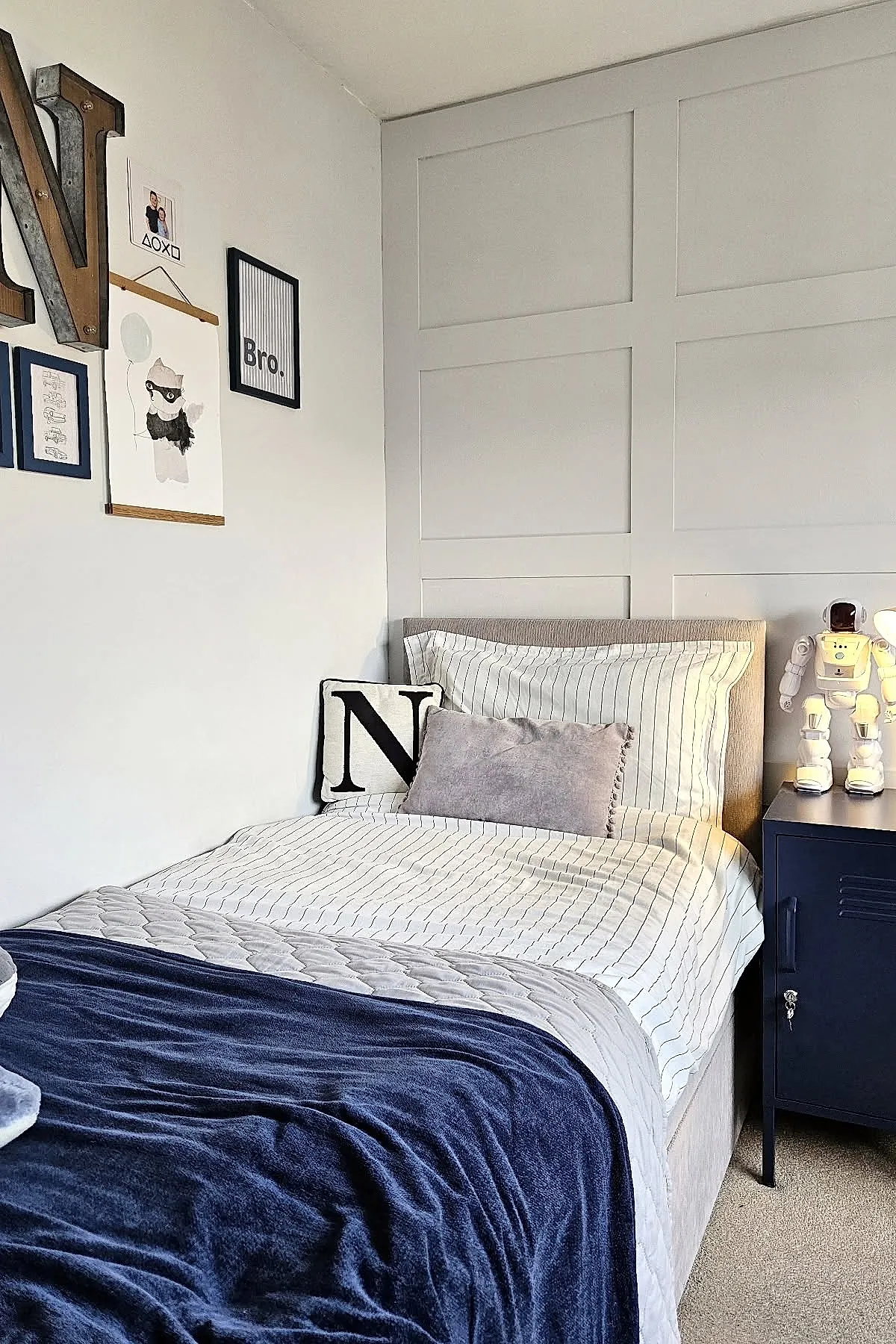 Teen boy bedroom featuring bed with white and black striped bedding, light gray decorative pillow, black and white letter N pillow, beige upholstered headboard, dark blue nightstand holding a white retro robot lamp against light gray paneled wall.