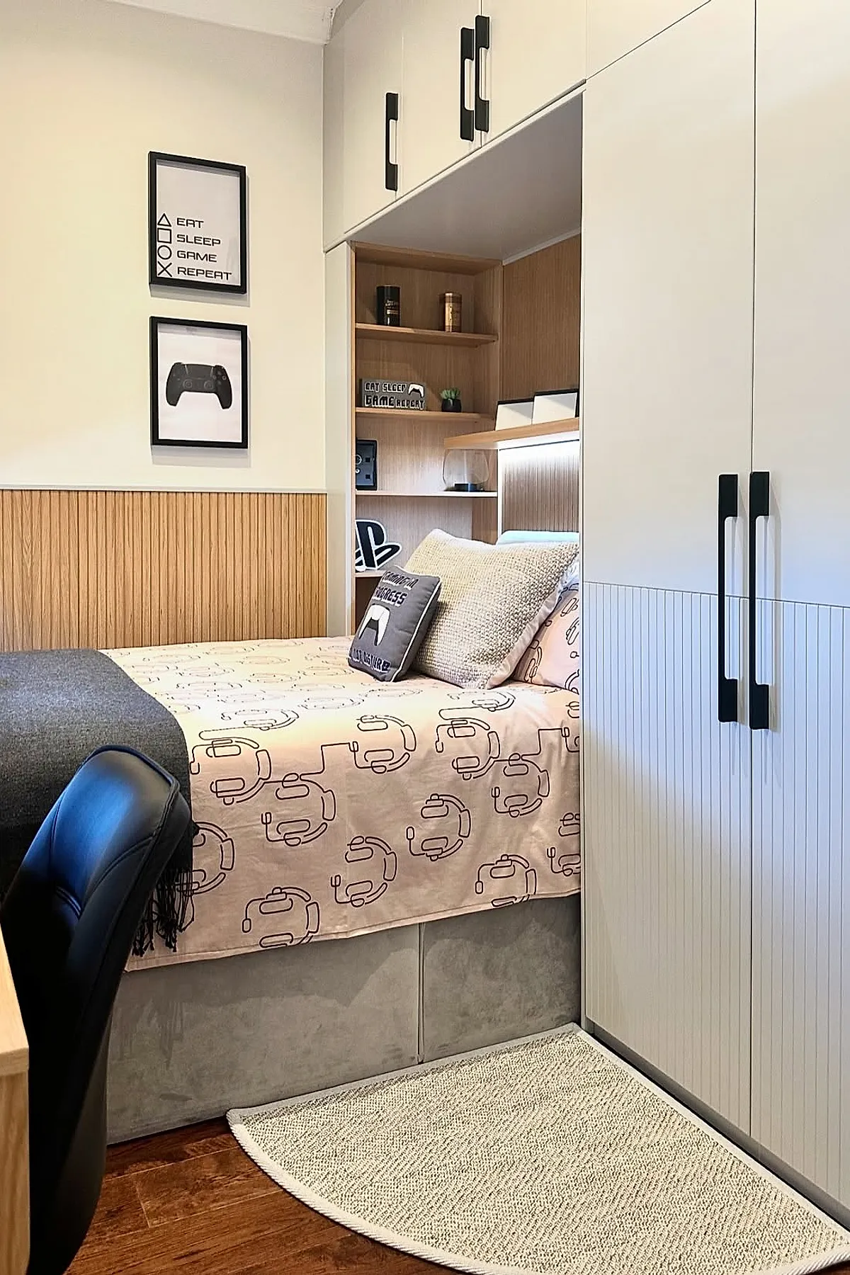Teen boy bedroom featuring a bed with headset-patterned pink bedding, beige textured pillow, built-in wooden shelves above headboard holding small decor items, white cabinets with black handles surrounding the bed, and framed gaming-themed prints on the wall.