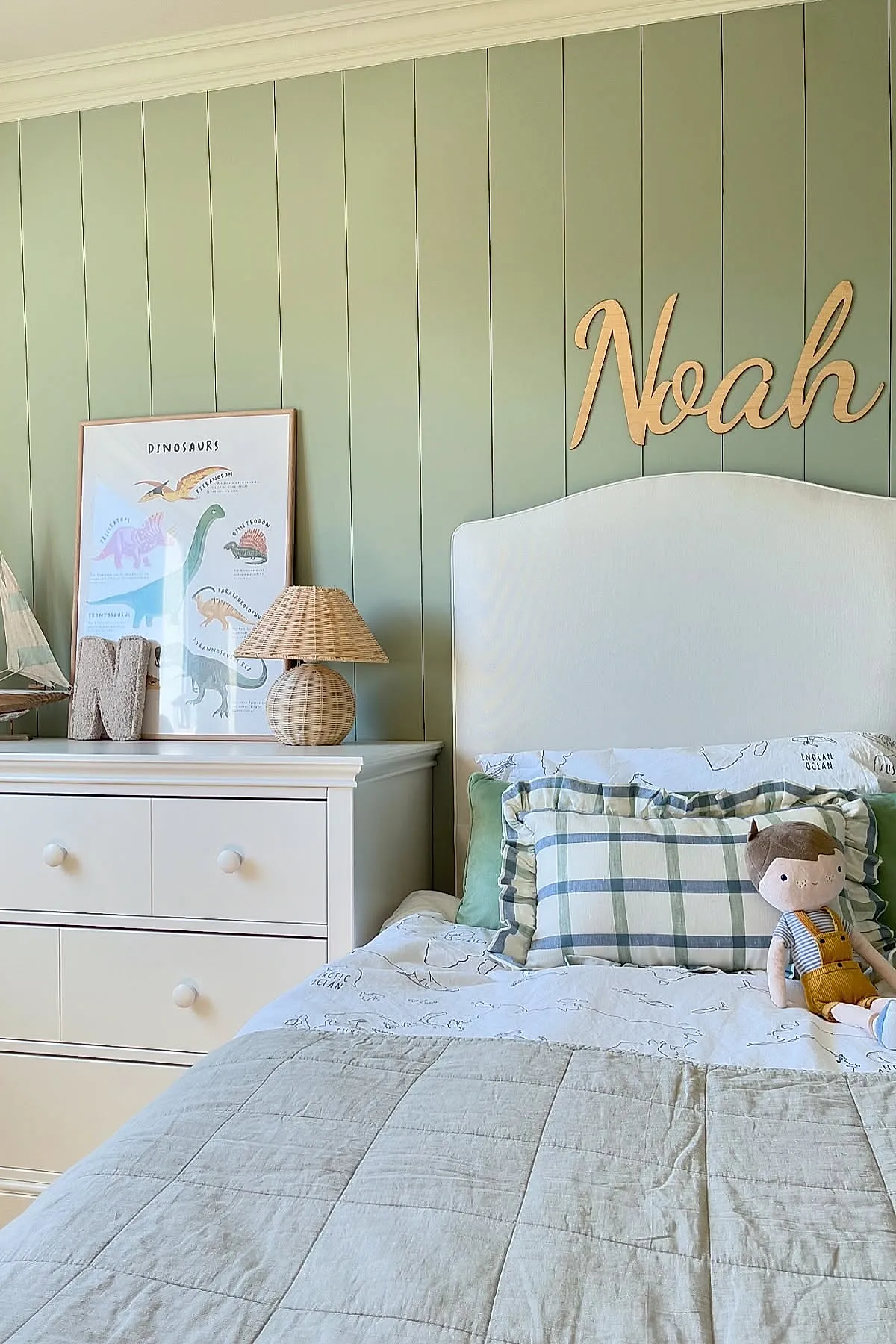 Boys bedroom with pale green vertical paneled wall, white upholstered bed with plaid pillows, wooden name sign spelling Noah above bed, white dresser with wicker lamp, dinosaur poster, and decorative sailboat.