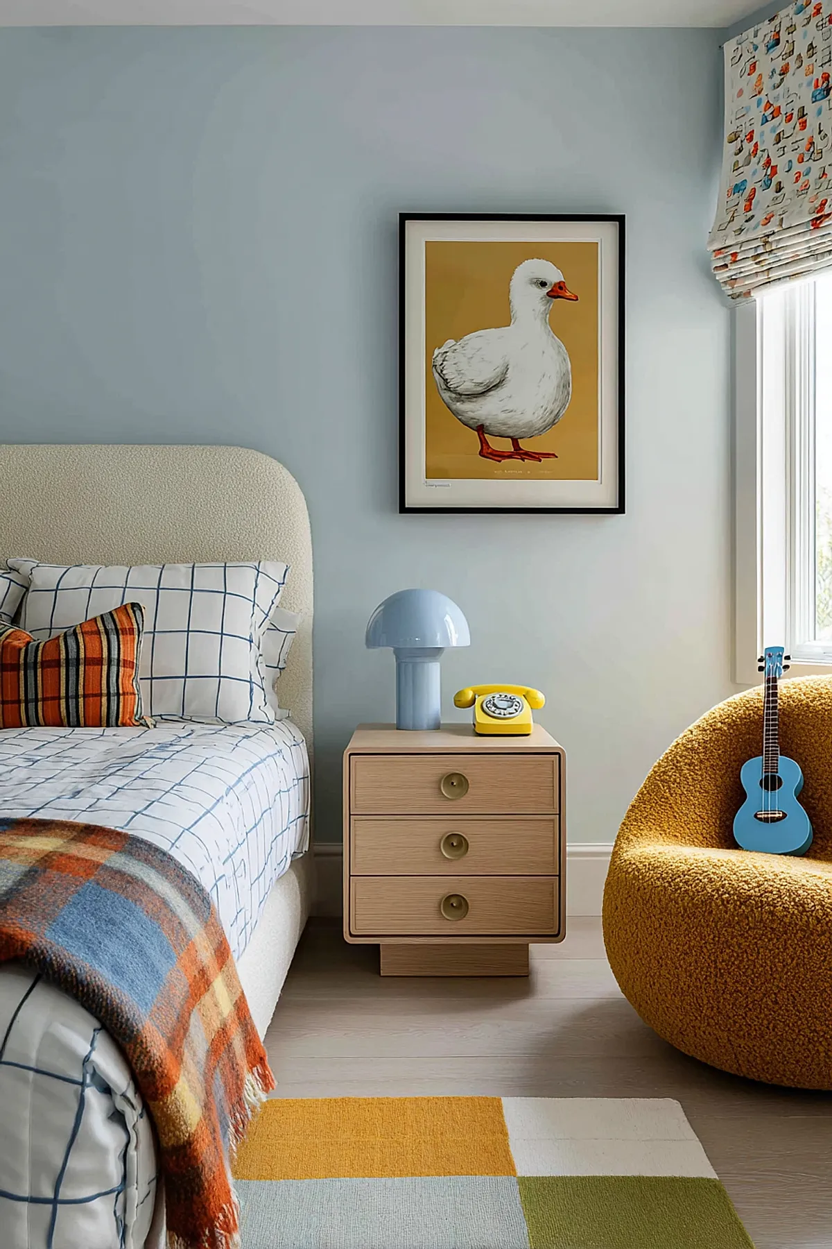 Boys bedroom featuring beige upholstered bed with checkered white and blue bedding, orange plaid throw blanket, light wood nightstand holding pale blue mushroom lamp and yellow rotary phone, framed print of white duck on mustard background, mustard yellow textured armchair with blue ukulele.