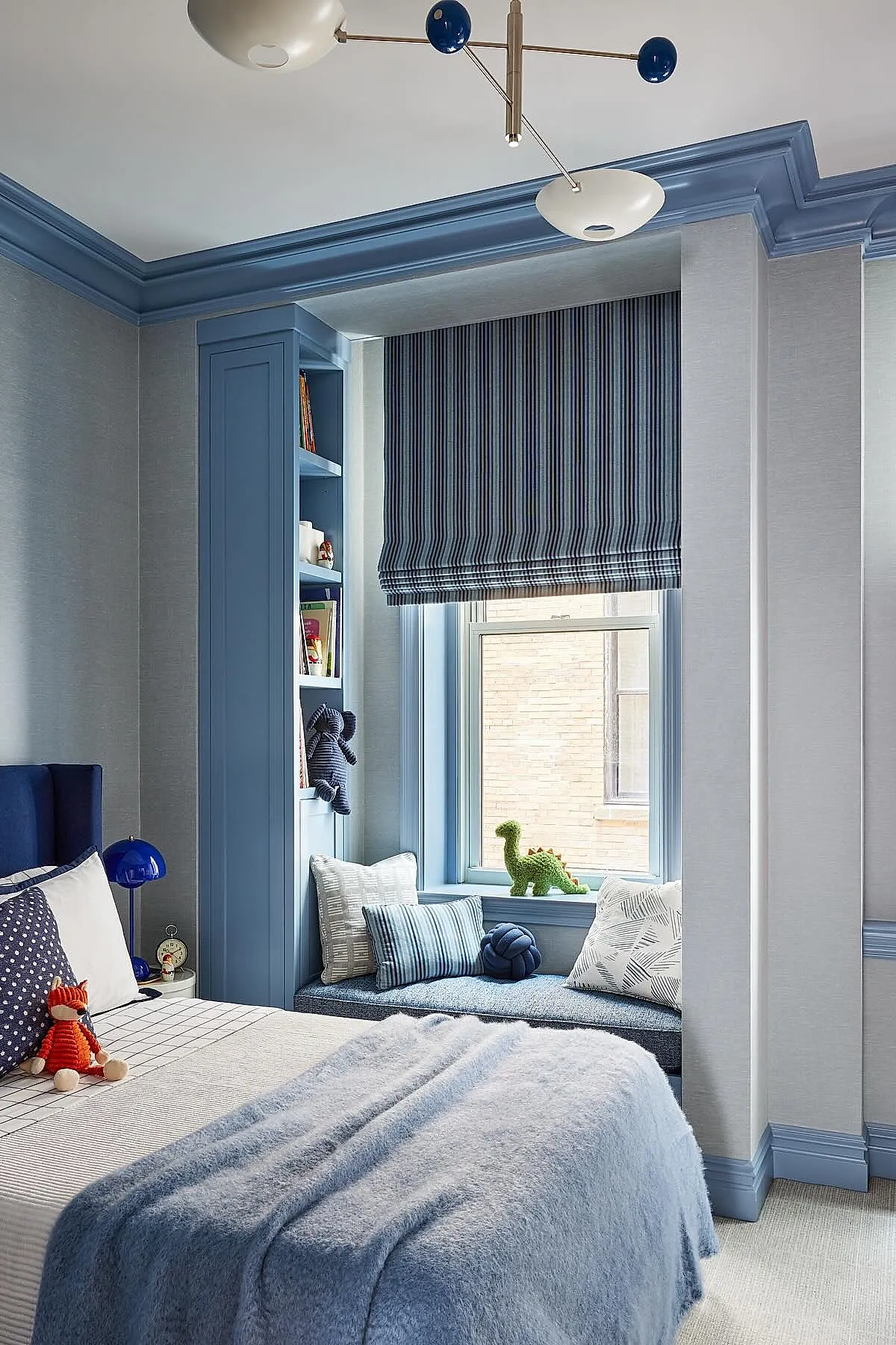 Boys bedroom featuring blue walls and ceiling trim, window seat with blue cushion and assorted pillows, striped blue Roman blind, bed with light bedding and plush toys including a fox, bedside table with blue lamp.