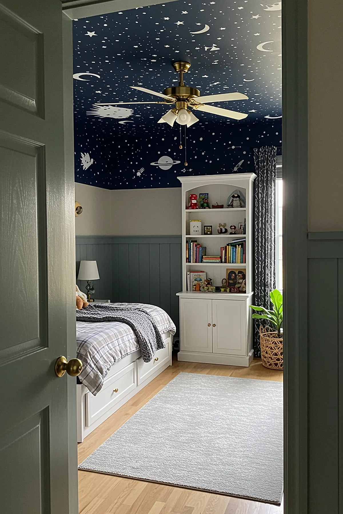 Boys bedroom with white bed and plaid bedding, blue-gray vertical wall paneling, white bookshelf filled with books and toys, deep navy ceiling painted with stars, moons, planets, gold ceiling fan.