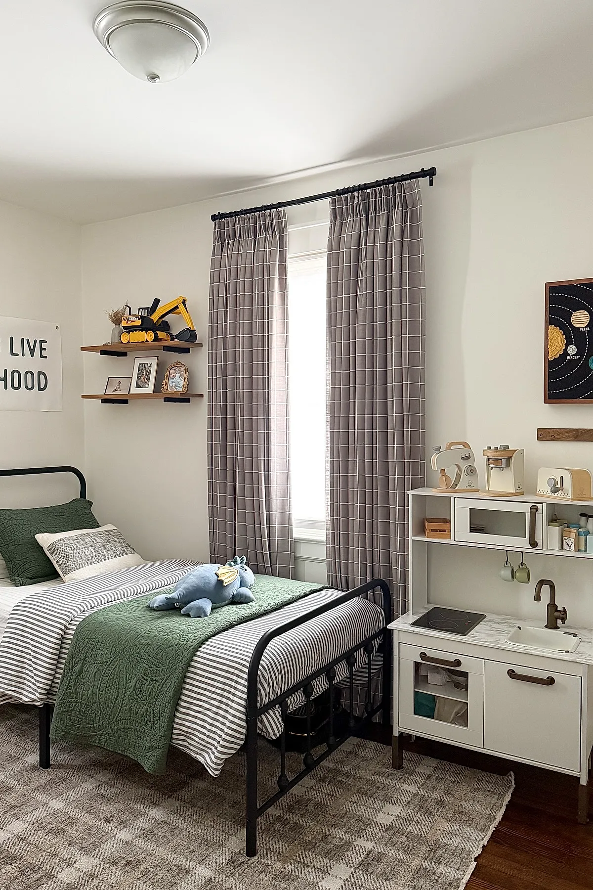 Boys bedroom featuring black metal twin bed with striped bedding and green quilt, gray grid patterned curtains, white play kitchen set, wooden shelves with toy excavator and framed photos, checkered beige rug on wood floor.