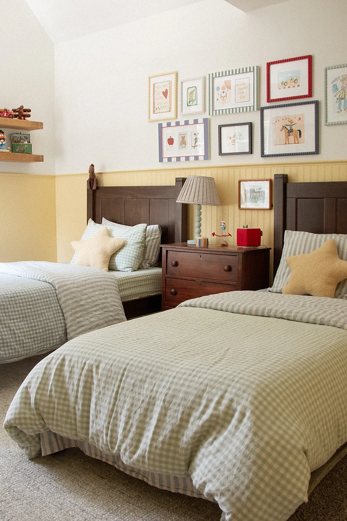 Boys bedroom with two dark wood twin beds dressed in pale green gingham bedding, cream star-shaped pillows, yellow wainscoting wall, framed kid art above beds, wooden nightstand with lamp.