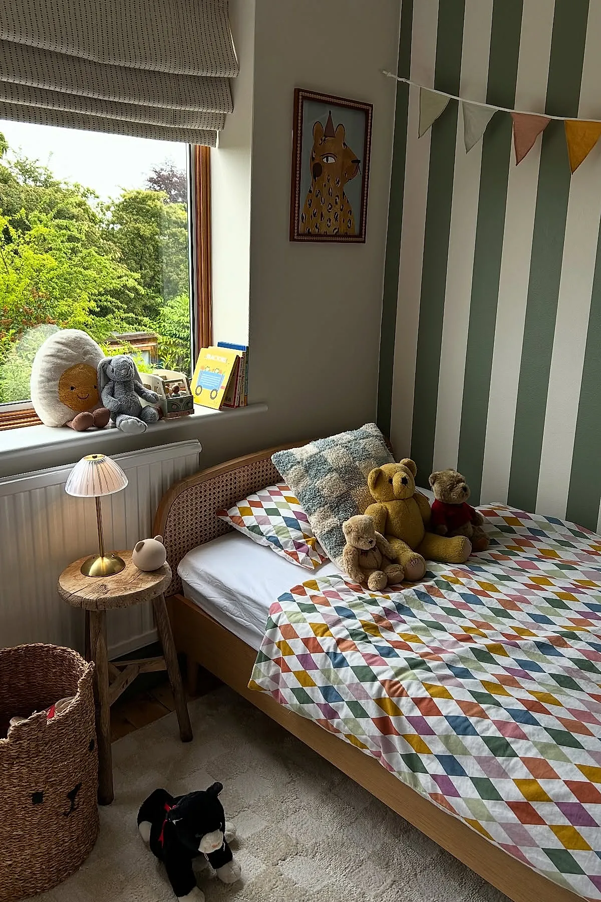 Boys bedroom with light wood bed featuring colorful diamond pattern bedding, green and white vertical striped accent wall, plush teddy bears on bed, rustic wooden side table with brass lamp, window seat with stuffed toys.