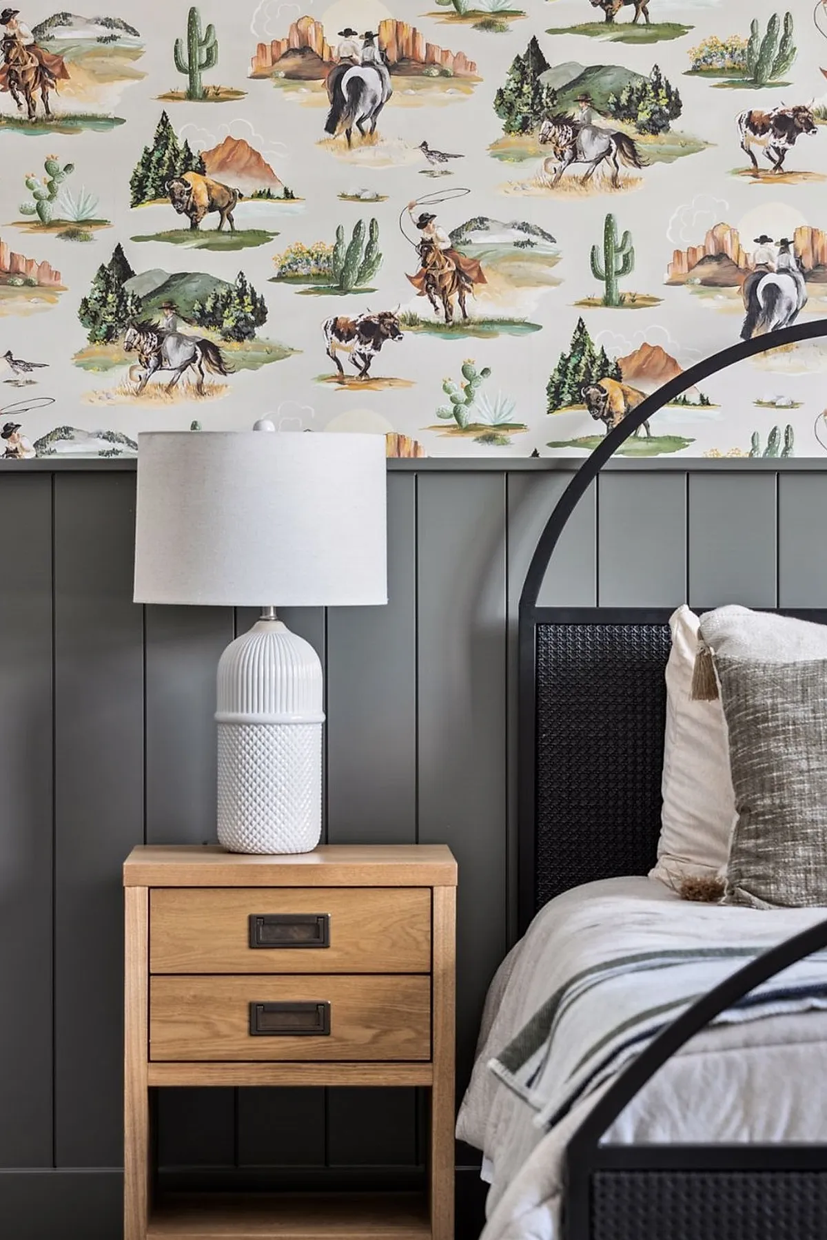 Close-up of boys bedroom featuring western-themed wallpaper with cowboys, horses, buffalo, cacti over gray vertical panel wall, wooden nightstand with white textured ceramic lamp beside black metal bed frame.