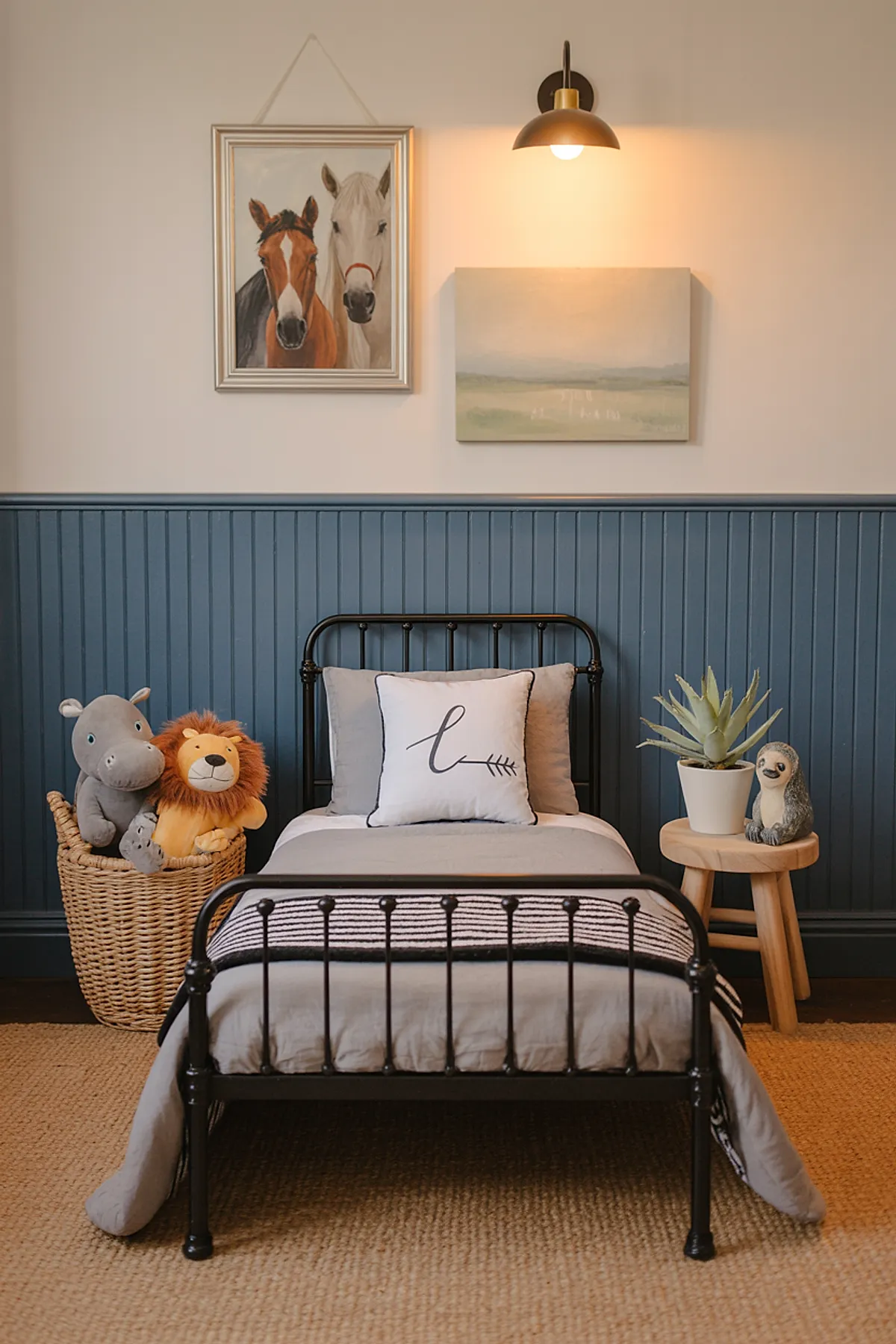 Boys bedroom featuring black metal bed with gray bedding, deep blue vertical wainscoting on wall, wicker basket with lion and hippo stuffed animals, small wooden stool holding a potted succulent and sloth figurine, framed paintings of horses and landscape above bed.