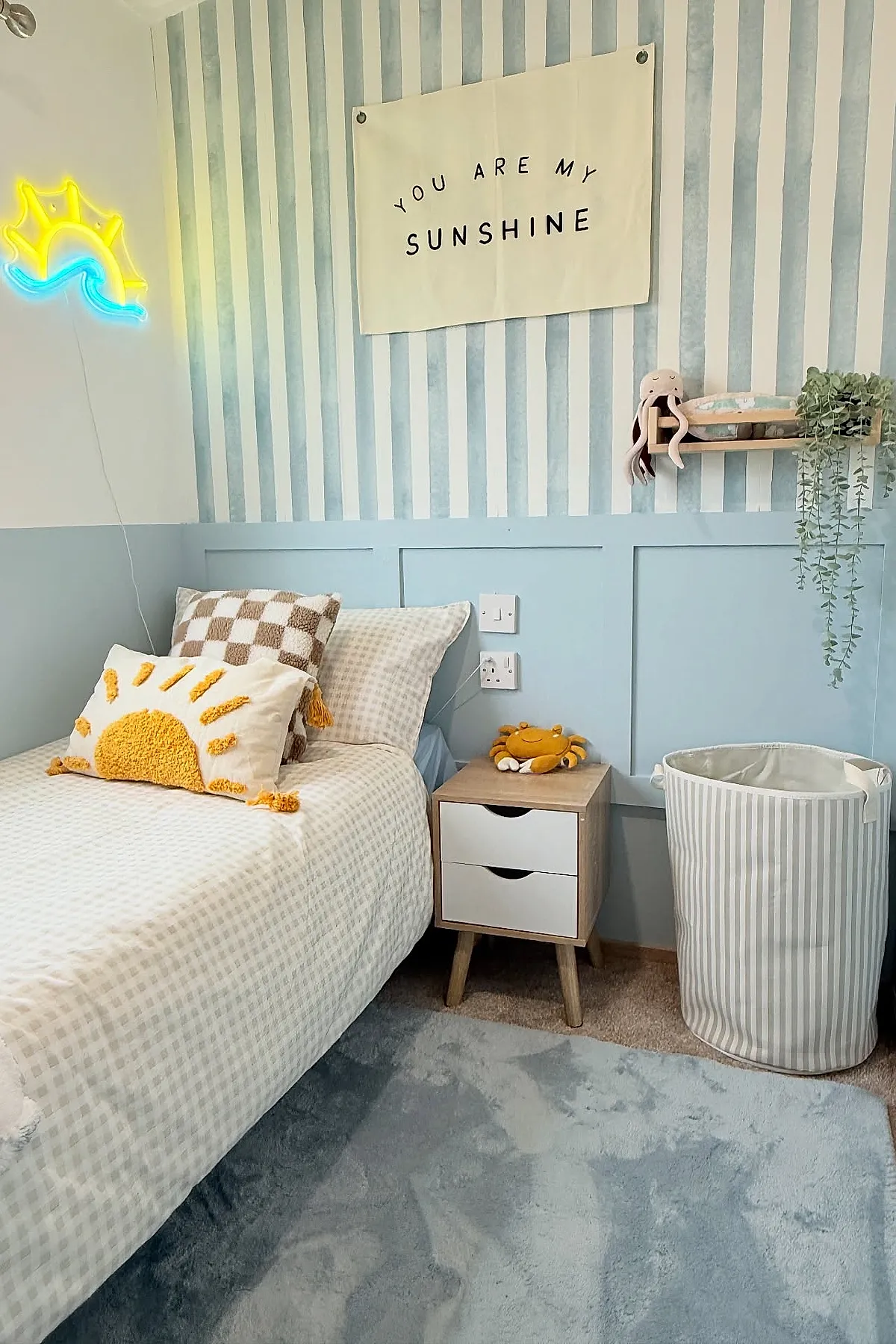 Boys bedroom with pale blue vertical striped wallpaper, half-painted blue panel walls, single bed with cream checked bedding and sun-shaped pillow, wooden nightstand with plush crab toy, neon yellow and blue sun wall light, white striped fabric laundry basket.