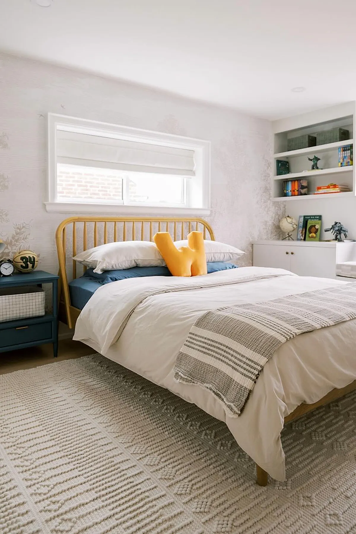 Boys bedroom with light wood bed frame, cream bedding, bright yellow U-shaped pillow, blue nightstand holding clock and toy ball, built-in white shelves with books and toys.