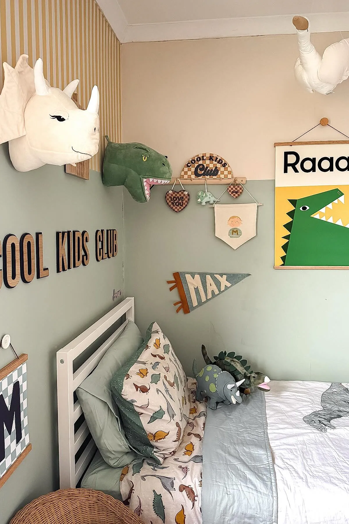 Boys bedroom with white bed frame, dinosaur print bedding, pale green walls half painted, stuffed dinosaur toys on bed, plush triceratops and T-Rex wall mounts, wooden 'Cool Kids Club' sign and Max pennant on wall.