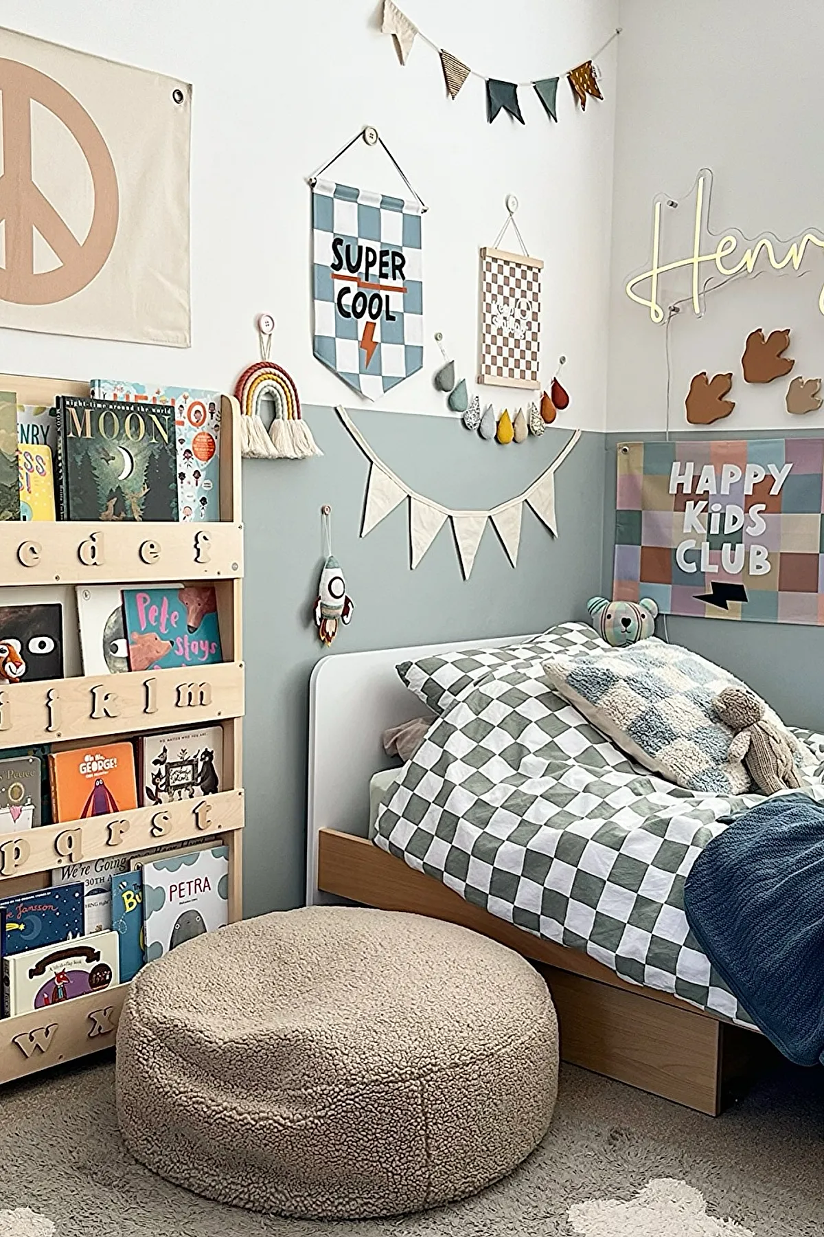 Boys bedroom featuring white and wood bed with green and white checkerboard bedding, pastel blue half-painted walls, colorful fabric banners and signs on wall, wooden bookshelf filled with books, beige textured round pouf on carpeted floor.