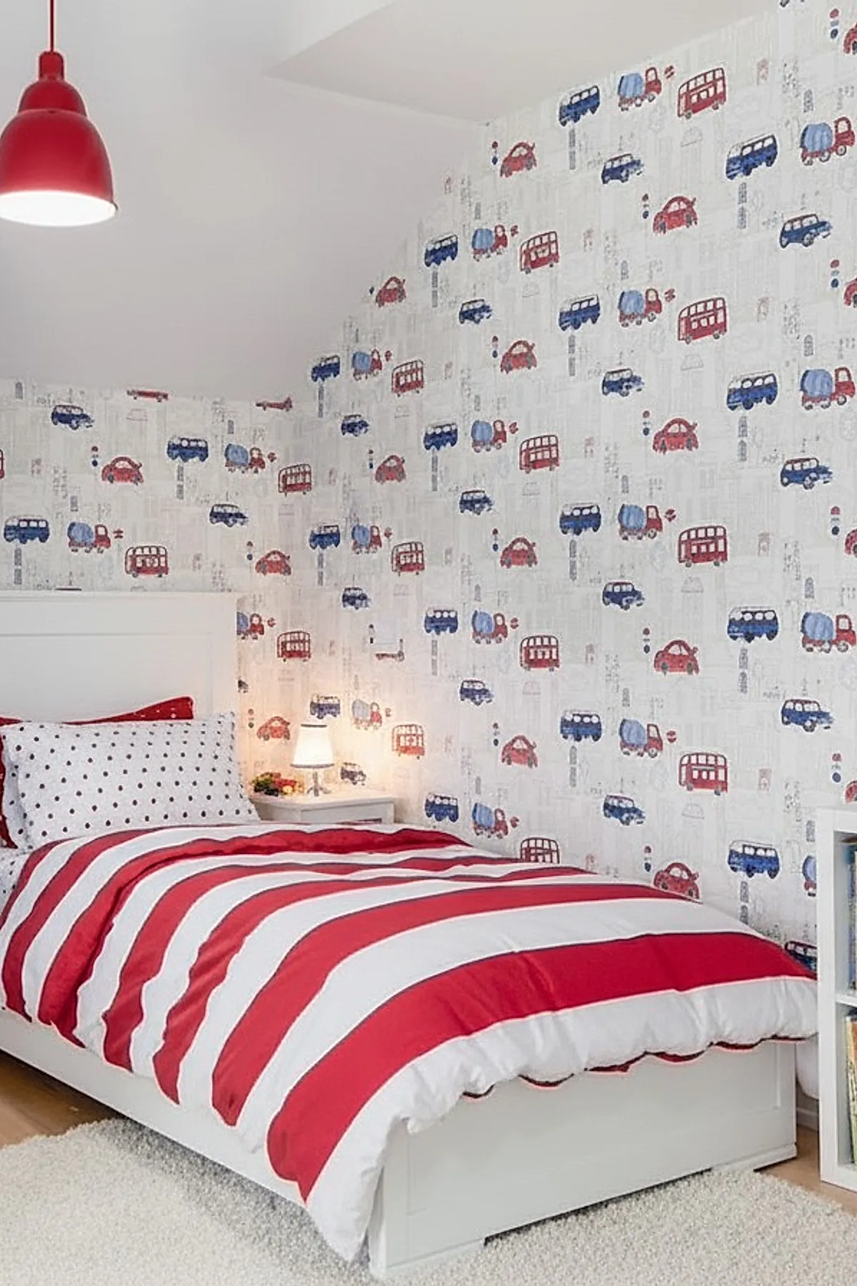 Boys bedroom with white bedframe, red and white striped comforter, polka dot pillows, wallpaper featuring red and blue cars and buses, red hanging ceiling lamp.