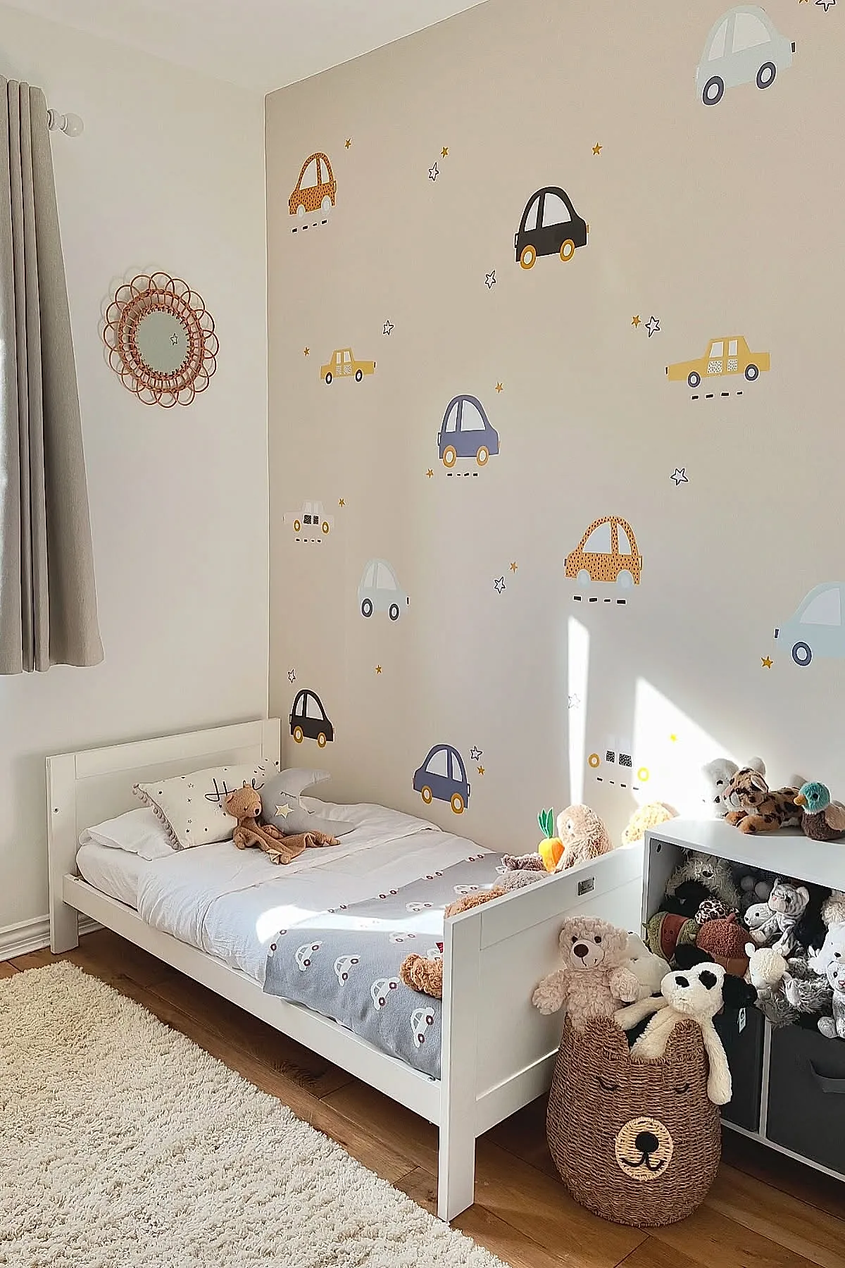 Boys bedroom with white wooden bed, beige wall decorated with colorful car decals, gray curtains, wicker toy basket shaped like a bear filled with stuffed animals, cream shag rug on wood floor.