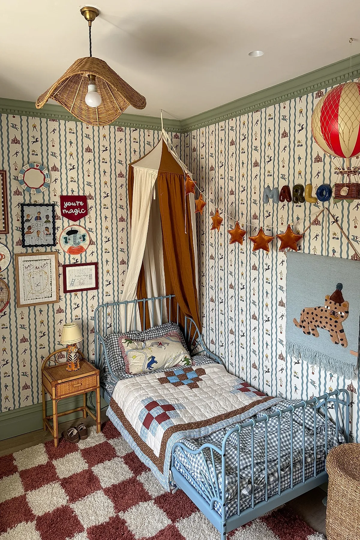 Boys bedroom featuring circus-themed wallpaper, blue metal bed frame with tent canopy, orange star garland, checkered red and white rug, wicker nightstand with tiger lamp.