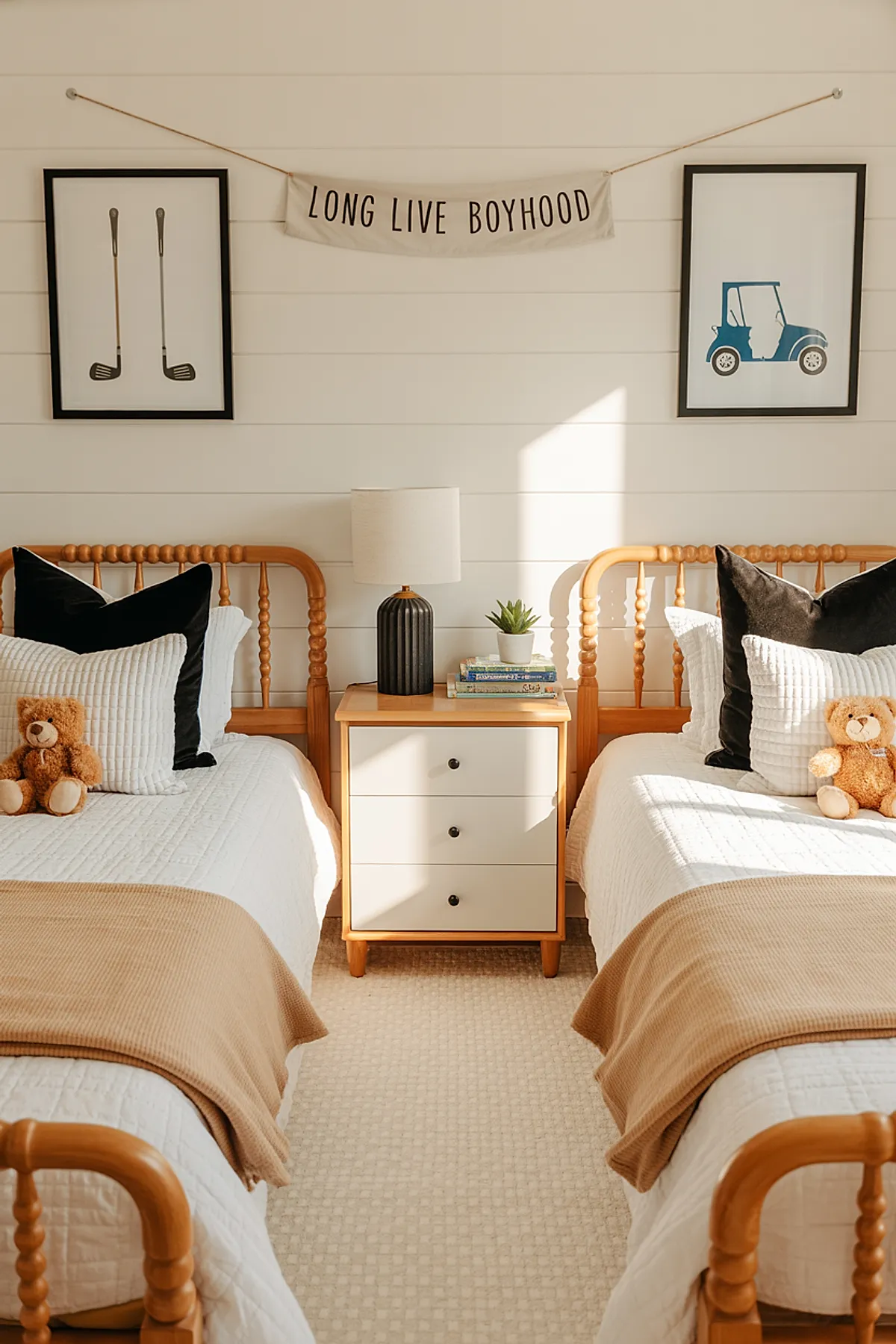 Twins bedroom with two wooden spindle twin beds dressed in white quilted bedding, black and white pillows, beige throw blankets, teddy bears on each bed, light wood nightstand with black lamp and small plant, wall art of golf clubs and blue golf cart with Long Live Boyhood banner between.