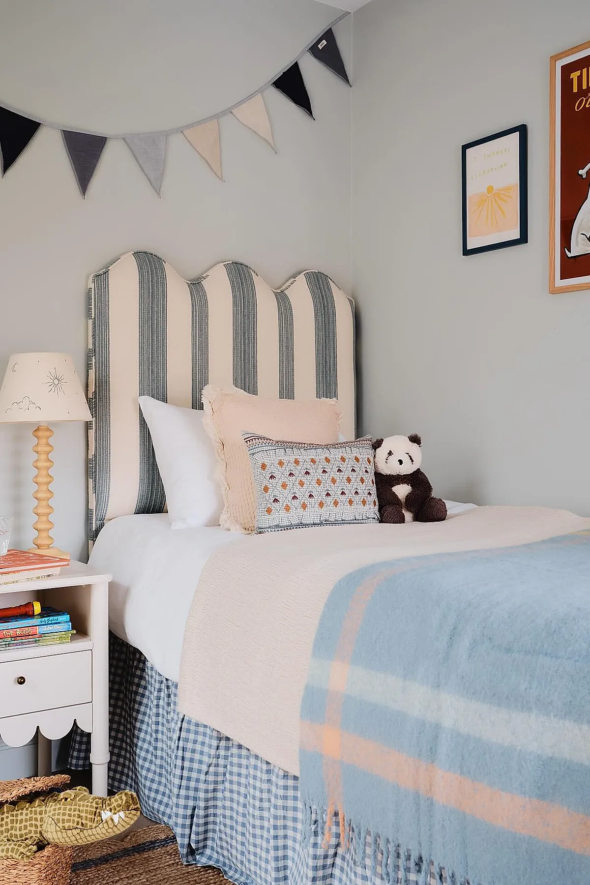 Boys bedroom featuring scalloped striped fabric headboard in cream and gray, white bedding with textured pastel pillows, plush panda toy, blue plaid bed skirt, light blue and peach throw blanket, gray walls with black white and beige pennant banner above bed.