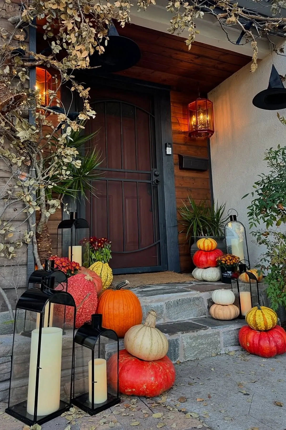various pumpkins in orange white yellow stacked on porch steps next to black metal lanterns containing large pillar candles under hanging dried branches