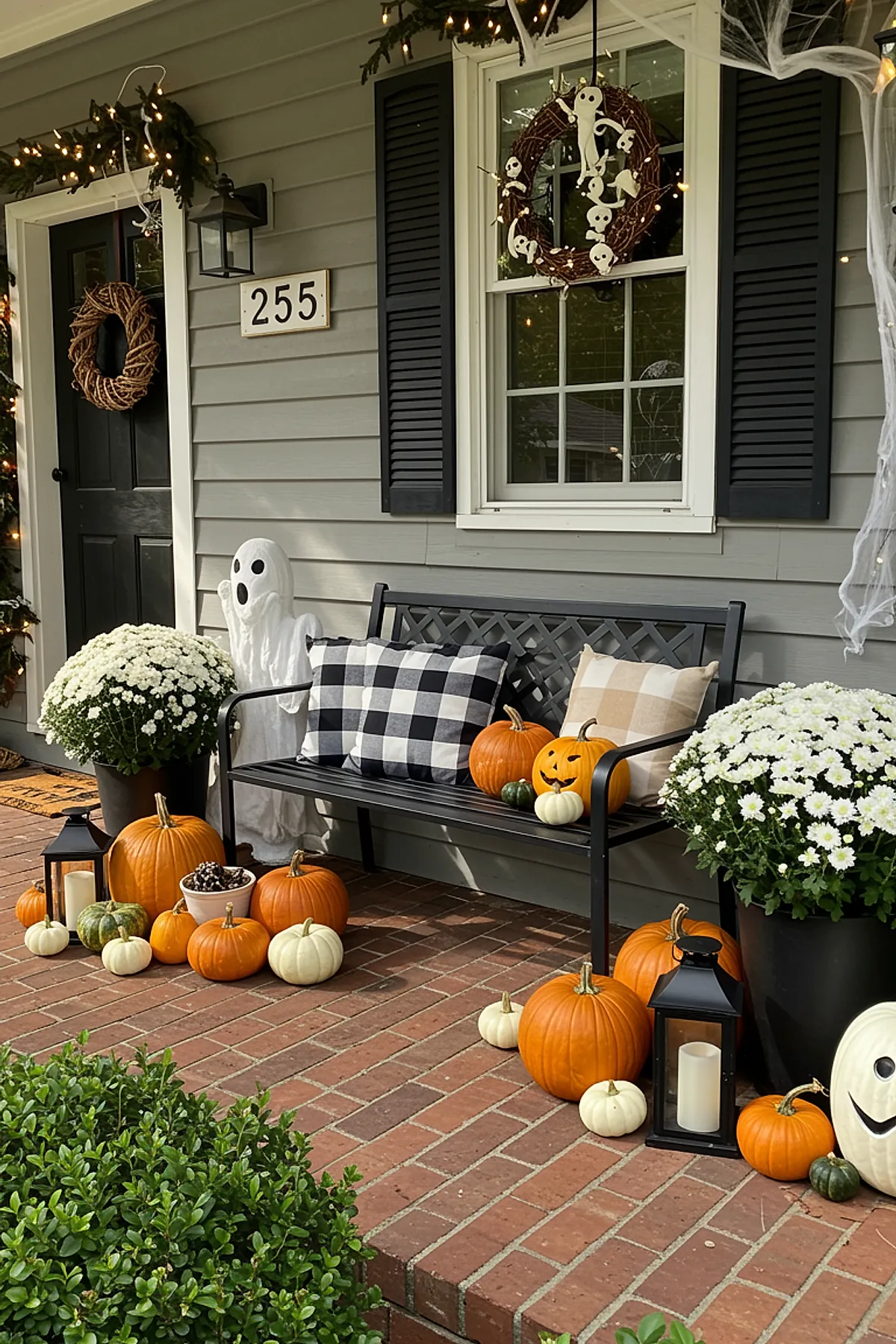 black metal bench on brick porch surrounded by assorted orange, white, and green pumpkins, two white ghost decorations, large pots of white chrysanthemums, black lanterns with candles, checkered black and beige pillows
