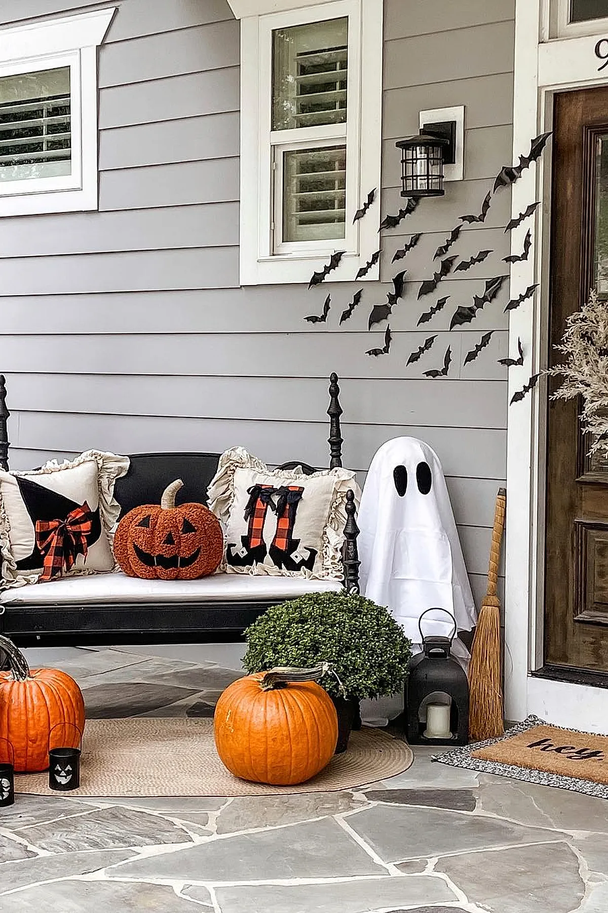 front porch with black bench holding jack-o-lantern pillow and witch legs pillow, white fabric ghost figure, orange pumpkins on stone floor, black lantern, wooden broom, cluster of black bat decorations flying up gray siding
