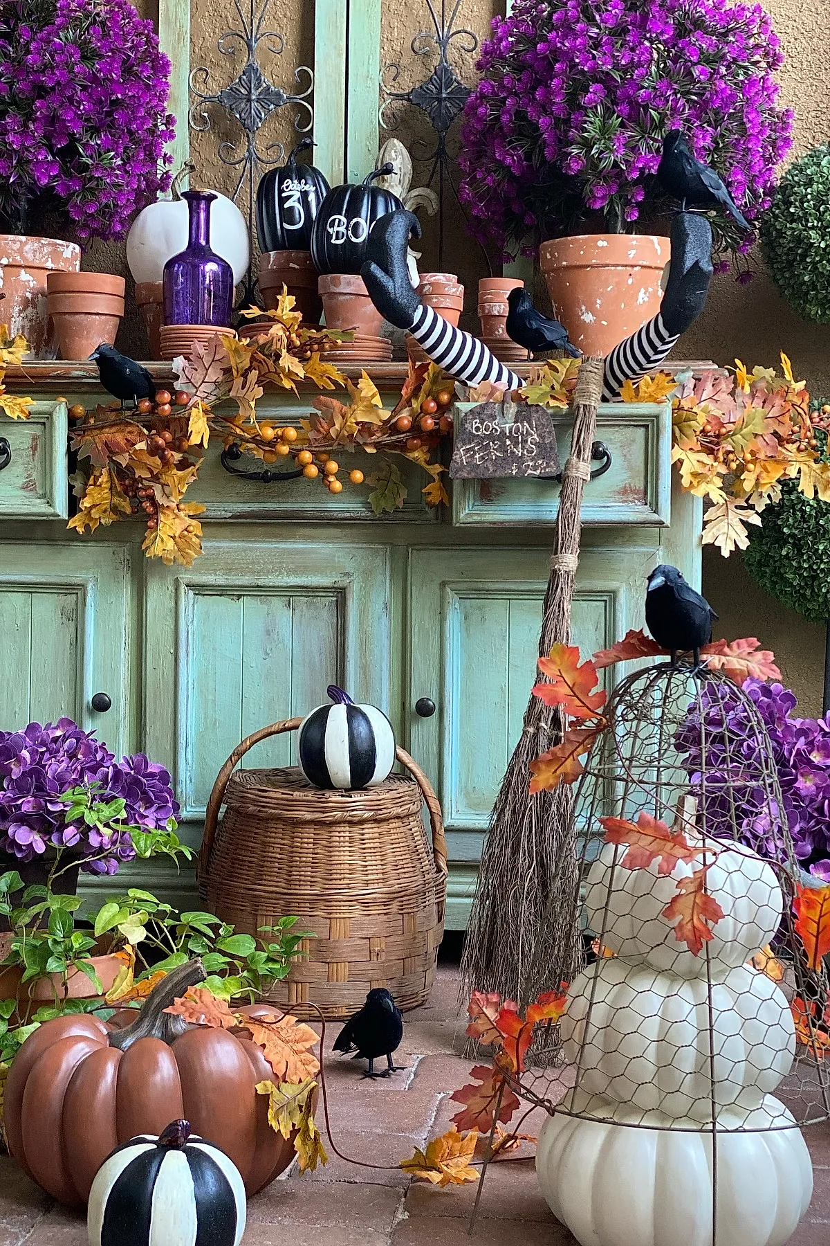 witch legs wearing black shoes and striped stockings sticking up from fall leaf garland surrounded by purple flowers terracotta pots black crows woven basket and pumpkins painted black white and brown
