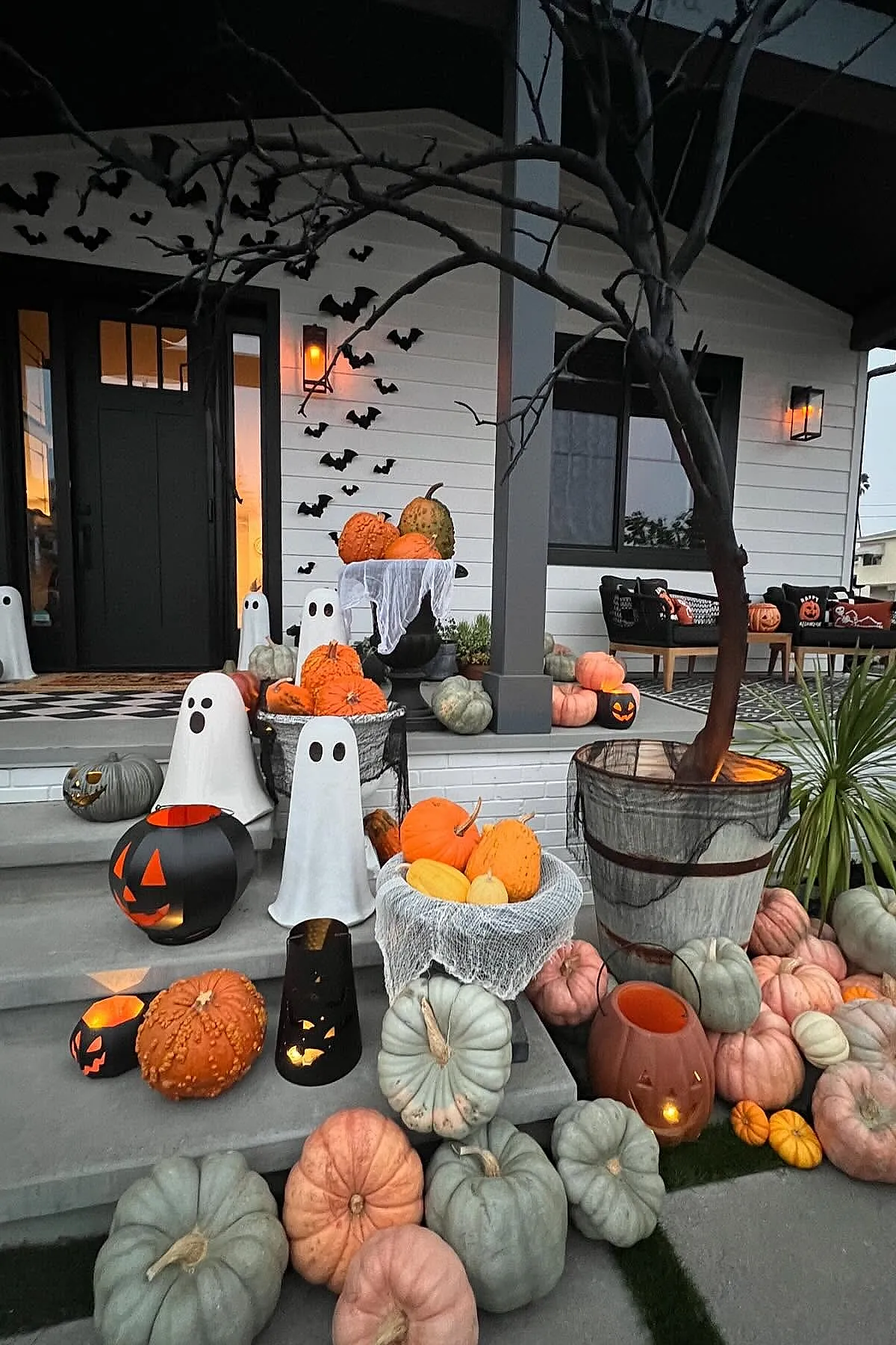 white ghost decorations with black eyes scattered pumpkins in shades of orange, gray, pink around steps, bare tree wrapped in black mesh fabric, black bat cutouts flying on white house exterior walls