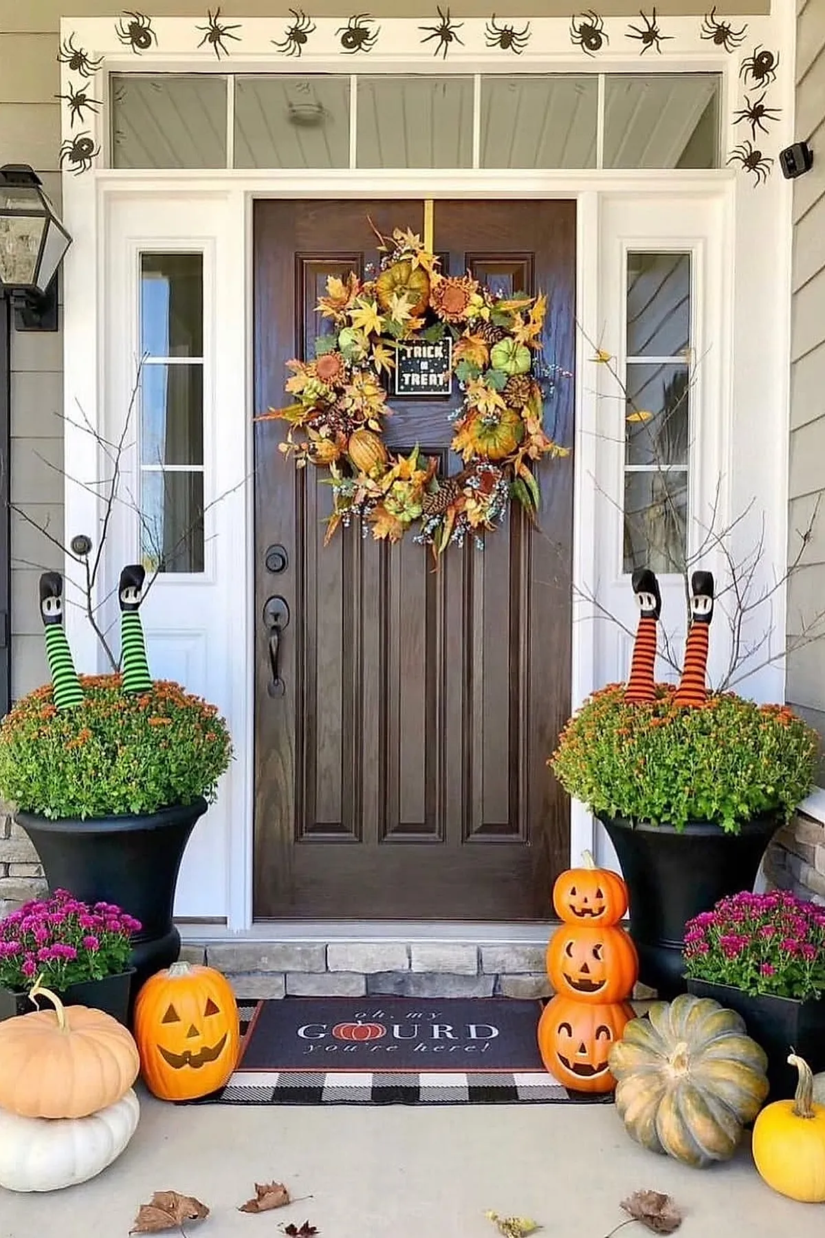 front porch decorated with two large black planters holding orange chrysanthemums with witch legs wearing striped stockings sticking out, brown wooden door adorned with autumn wreath, carved pumpkin jack-o-lanterns stacked and scattered, white pumpkins, and black spider stickers lining the door frame