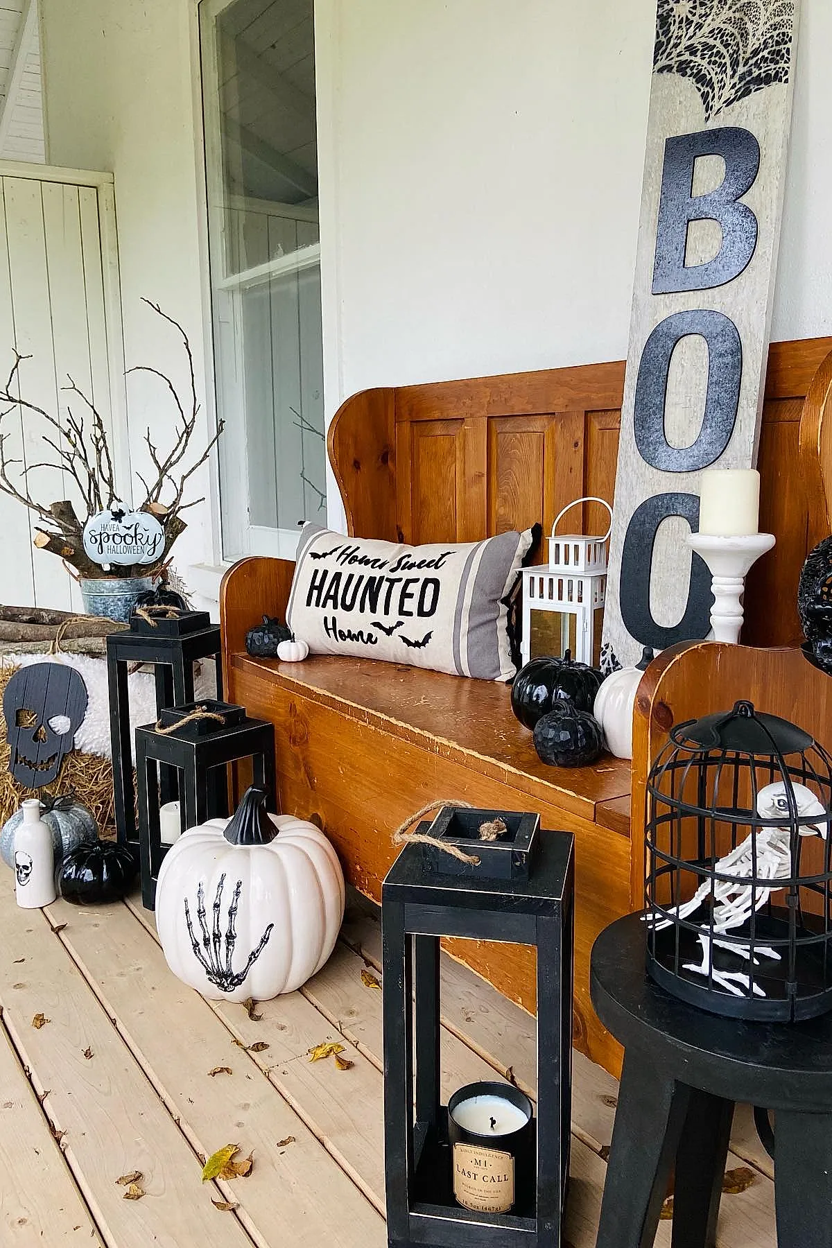 wooden bench with home sweet haunted home pillow surrounded by black and white pumpkins, black lanterns with candles, skeleton bird in black cage, skull decorated bottle, large wooden sign with BOO lettering