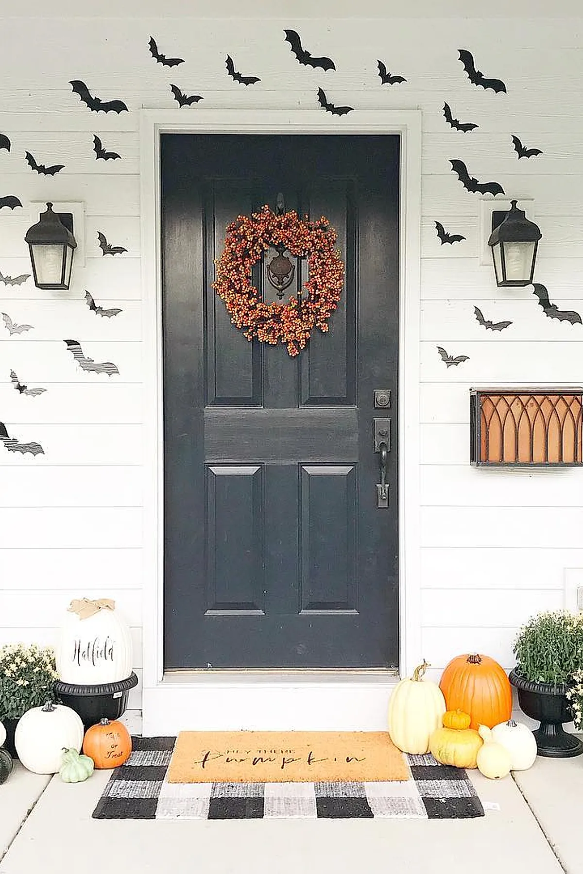 black front door with bright orange berry wreath surrounded by numerous black bat cutouts on white siding walls, assorted white and orange pumpkins grouped near potted mums on porch floor