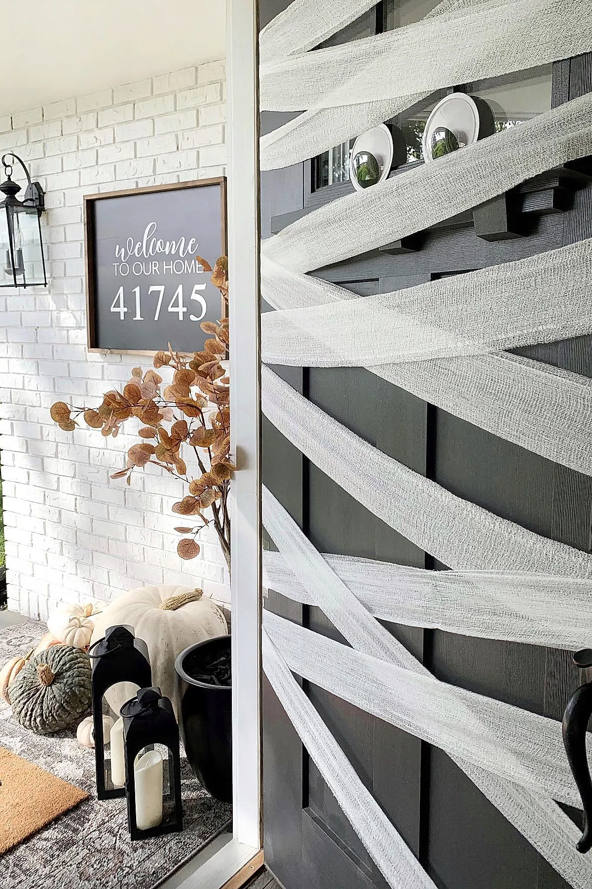 black front door wrapped in white gauzy fabric resembling mummy bandages with two large googly eyes attached, surrounded by white and green pumpkins, black lanterns with candles, dried brown branches in black planter