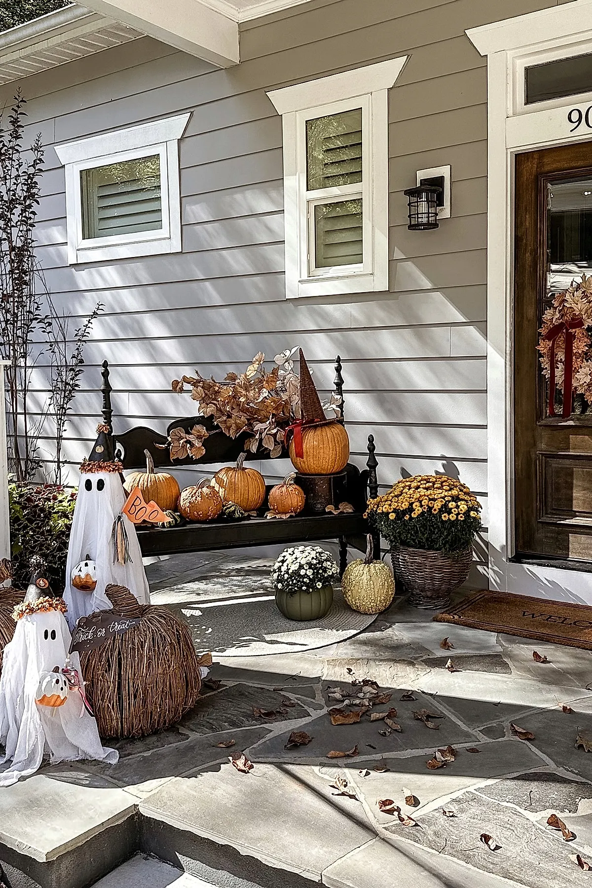 black wooden bench with orange pumpkins, pumpkin wearing witch hat, brown dried leaves behind, two white ghost figures with 'boo' sign, yellow and white potted mums on stone porch near dark wood door with autumn wreath