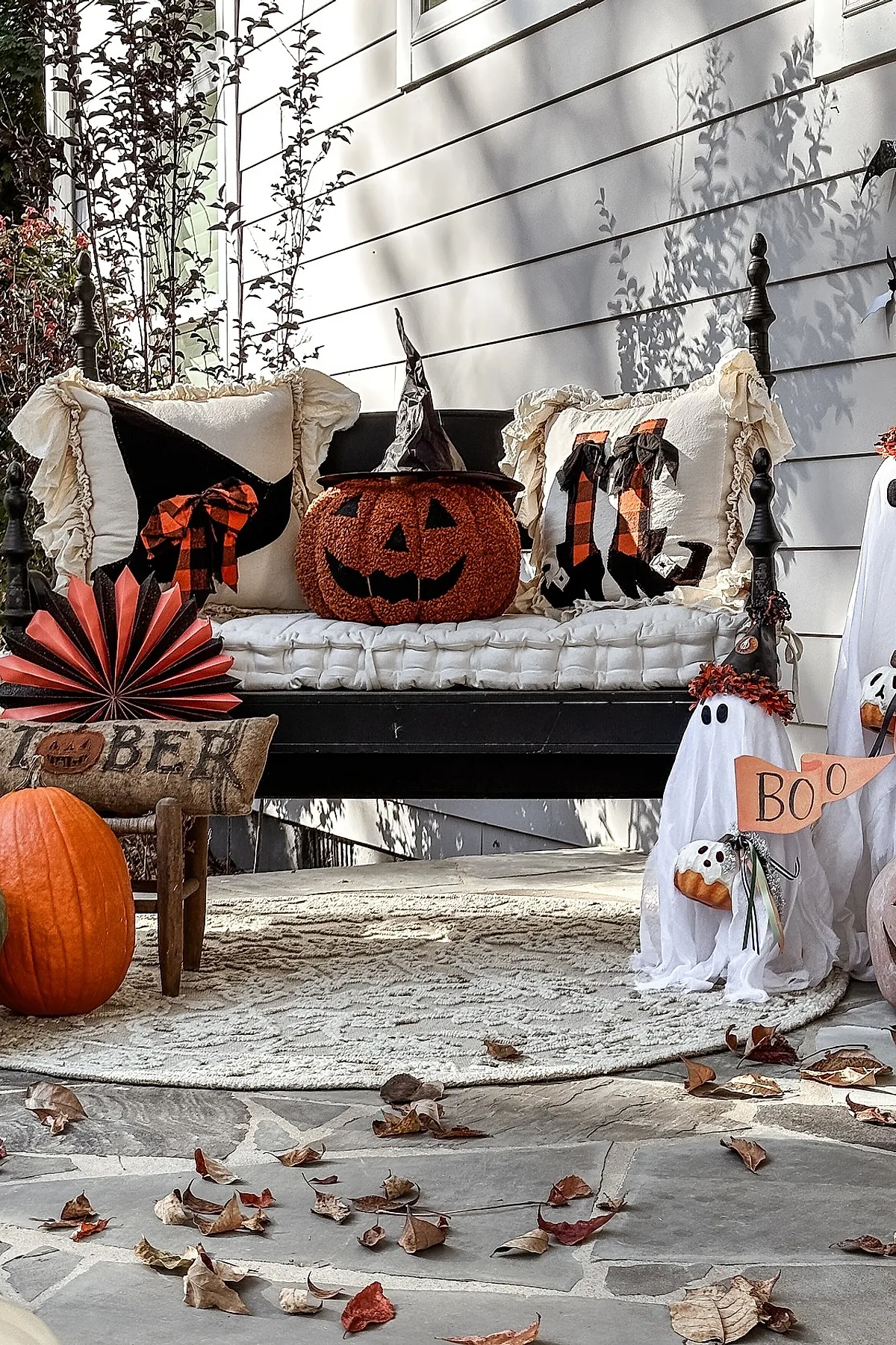 black wooden bench with white cushioned seat holding two ruffled edge pillows with Halloween designs and a textured pumpkin wearing a black witch hat, surrounded by small ghost decorations, orange pumpkin, and fall leaves scattered on gray stone floor