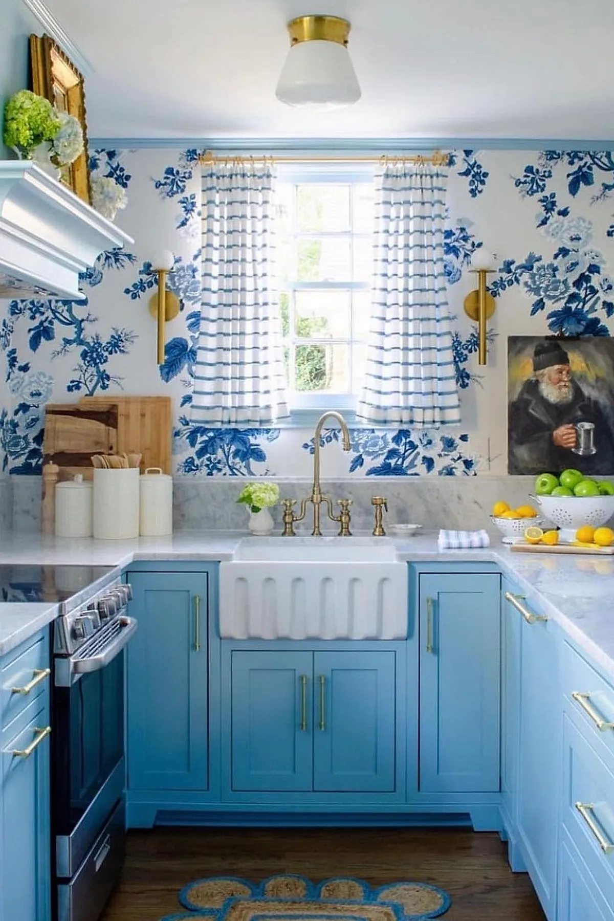 small kitchen with light blue cabinets, white farmhouse sink with brass faucet, blue and white floral wallpaper, striped curtains on window, brass wall sconces, marble countertops