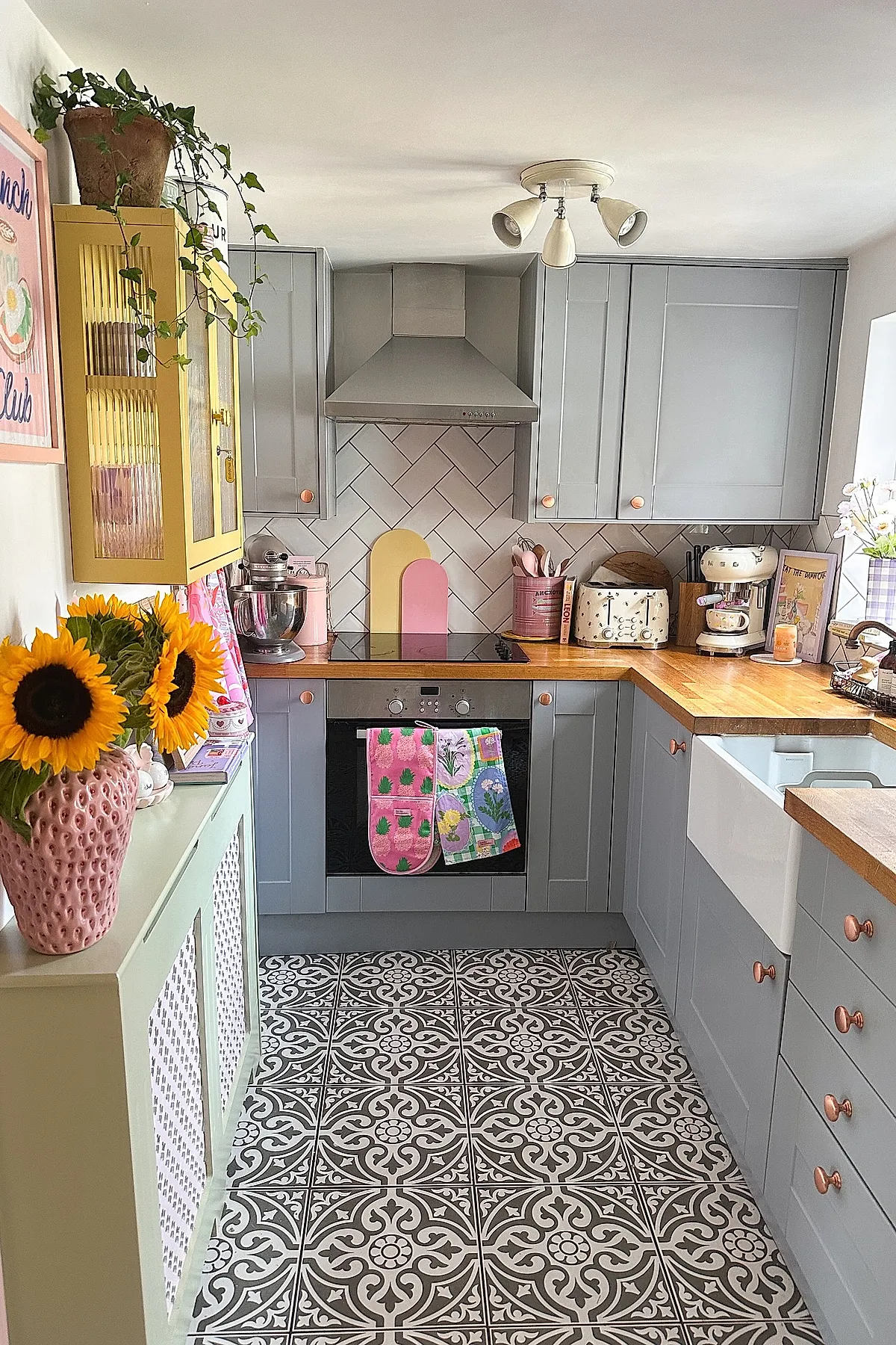 small kitchen with gray cabinetry, wooden countertops, black and white patterned floor tiles, yellow wall cabinet, sunflowers in pink vase, pink and pastel kitchen accessories