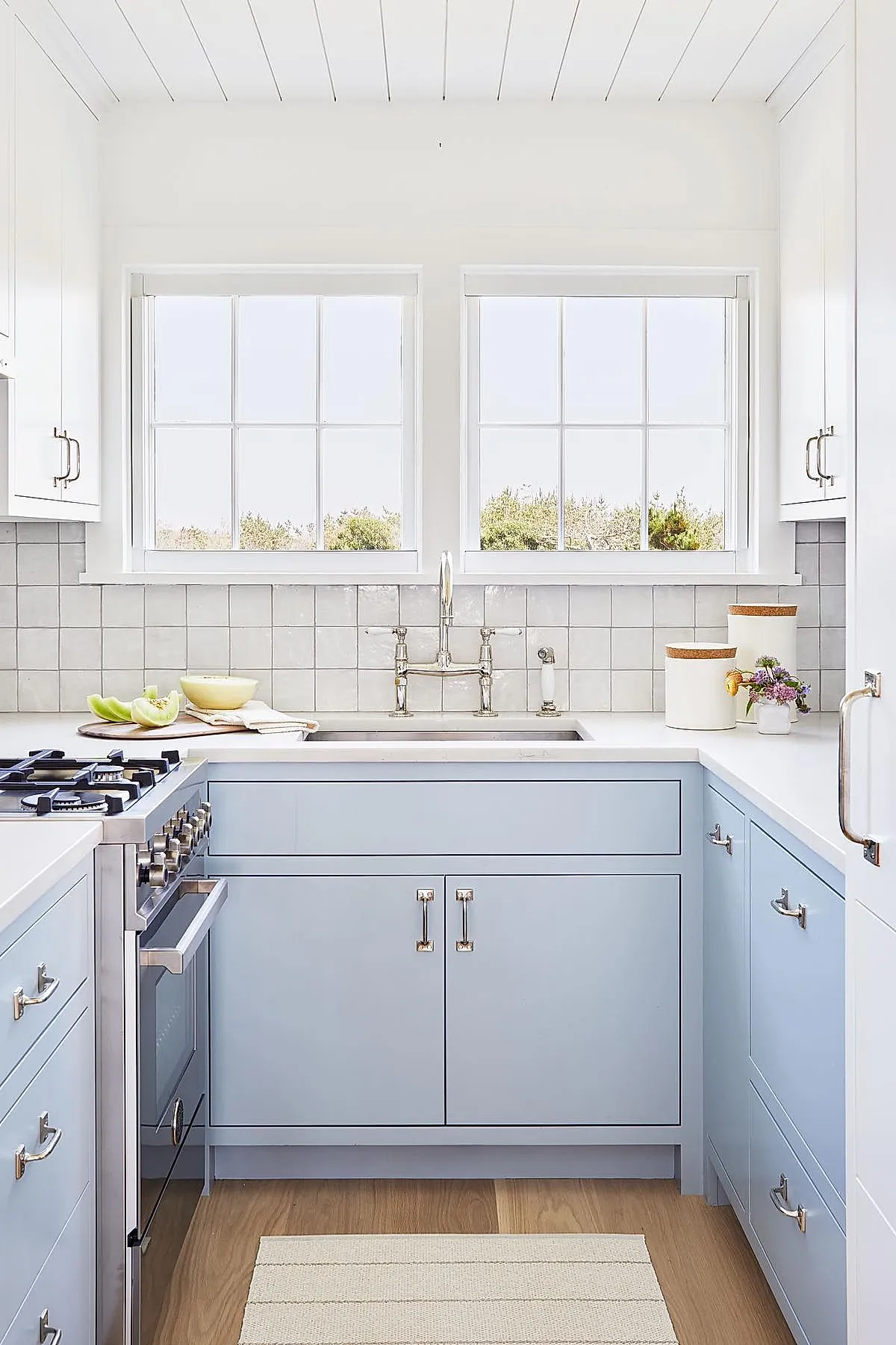 small U-shaped kitchen with light blue lower cabinets, white tiled backsplash, white countertops, stainless steel stove, two large windows above sink letting in natural light