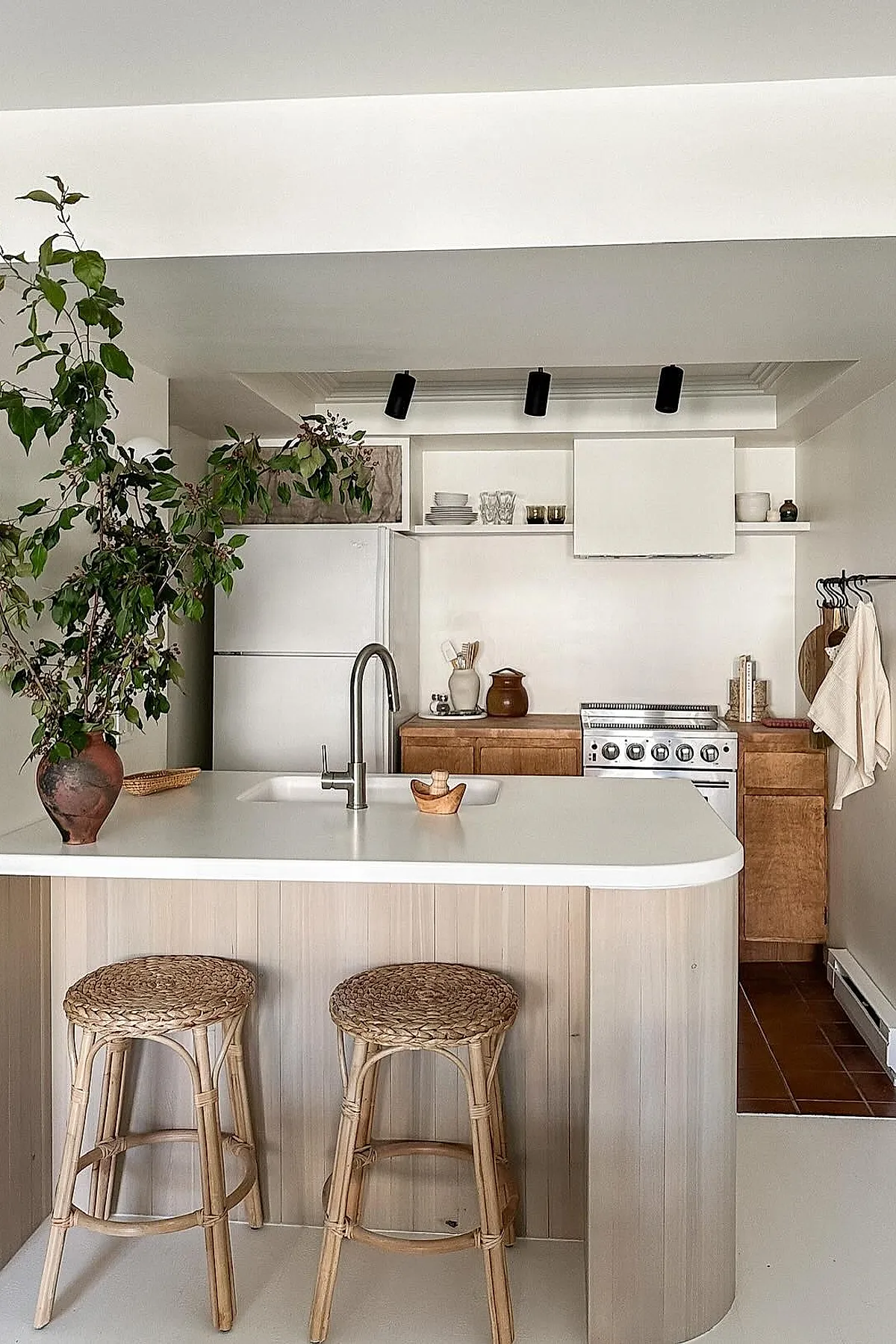 small open kitchen with light wood cabinets, white island countertop, stainless steel faucet, rattan stools, potted green plant on counter, black track lights