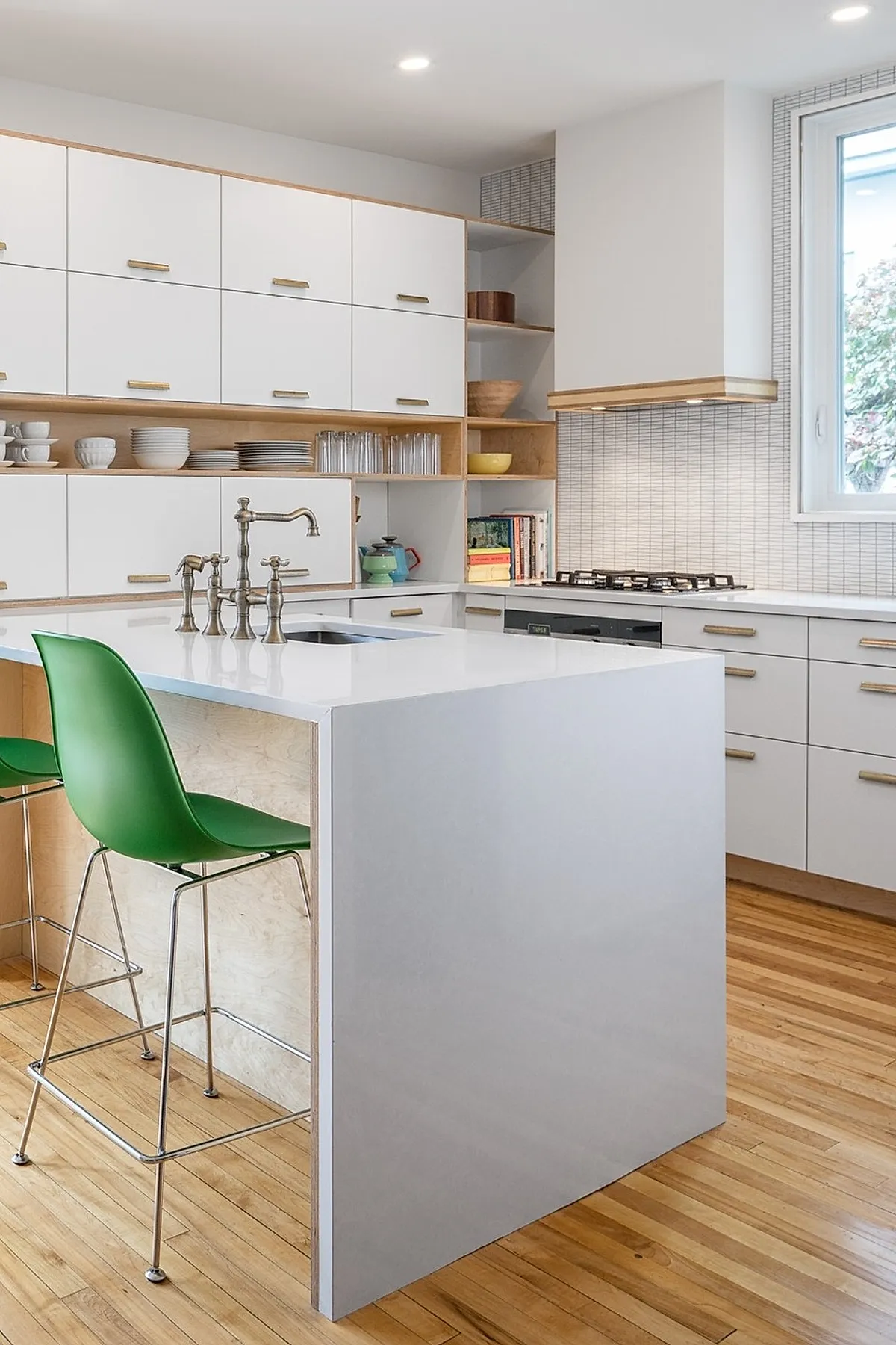 small kitchen with white cabinetry and island, brass handles, green plastic bar stools with metal legs, light wood flooring, stainless steel faucet, window by stove area
