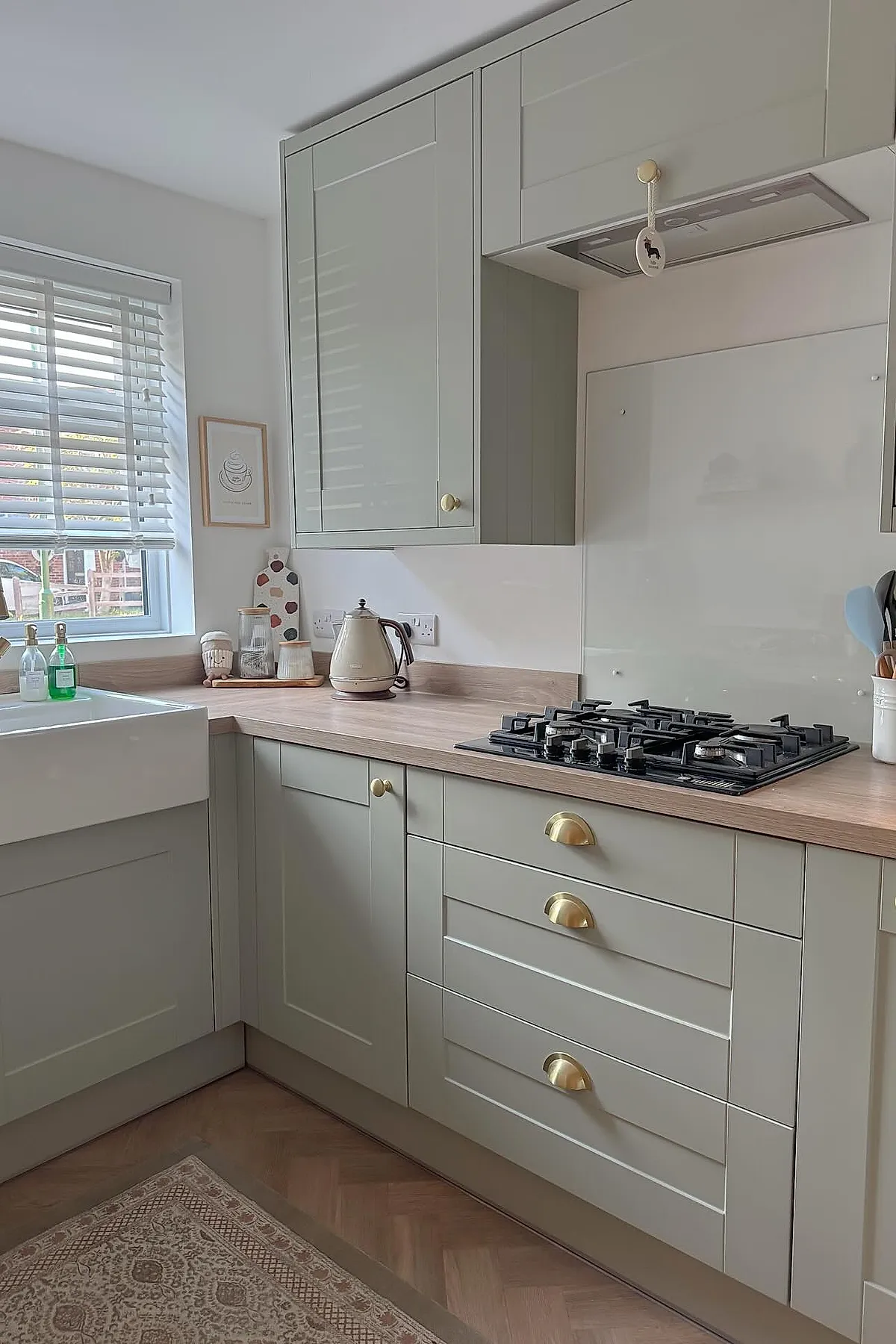 small kitchen featuring light gray cabinetry, brass handles, wood countertops, white farmhouse sink under window with blinds, gas stove with black grates, neutral walls