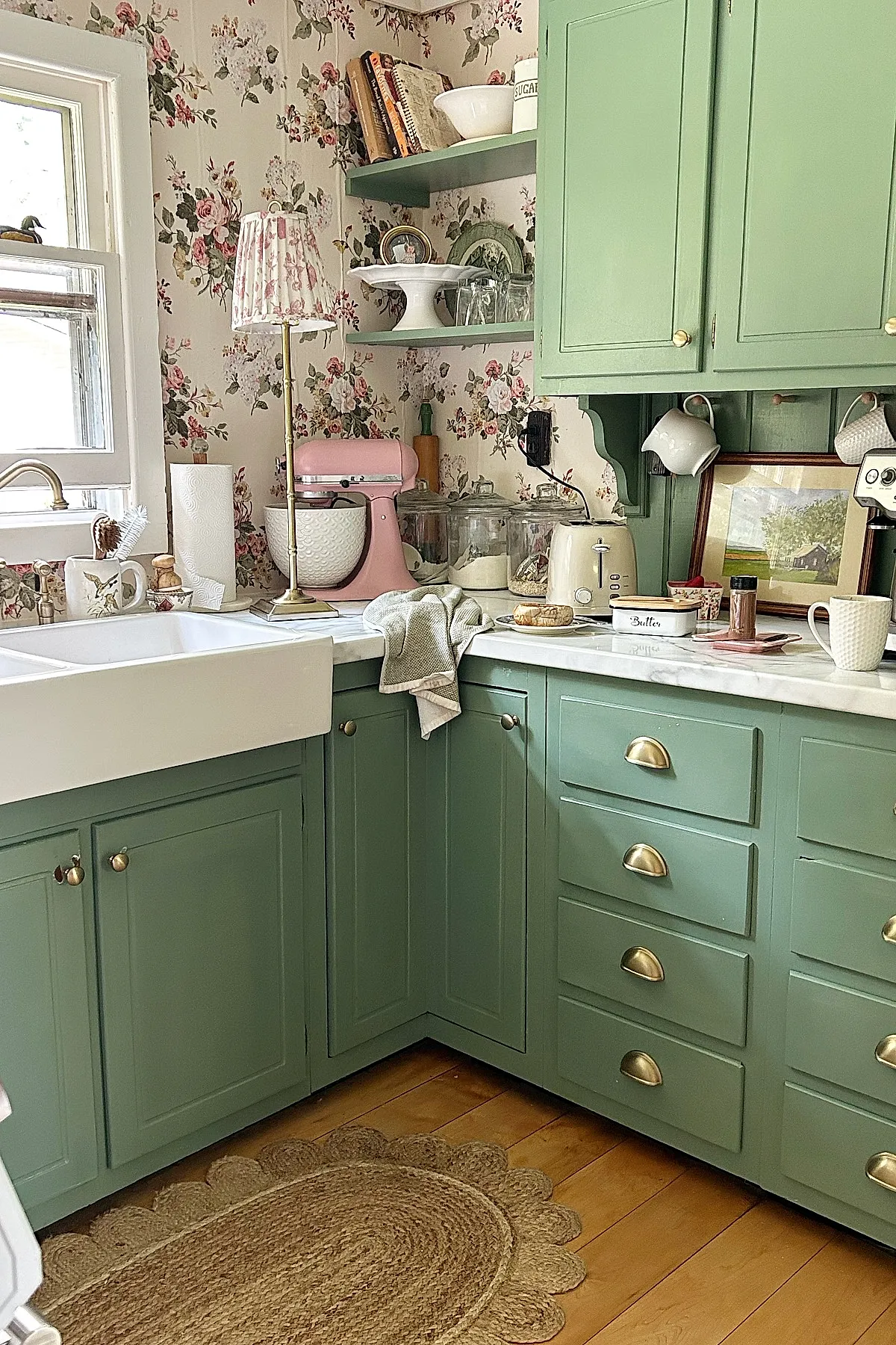 small kitchen with pastel green cabinets, floral wallpaper, white farmhouse sink, pink stand mixer, brass hardware, marble countertops, scalloped jute rug on wood floor