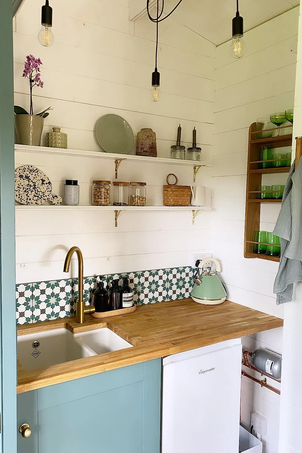 small kitchen featuring teal lower cabinets, wood countertop with inset white sink, brass faucet, green and white patterned tile backsplash, open shelves with jars and dishes, hanging light bulbs